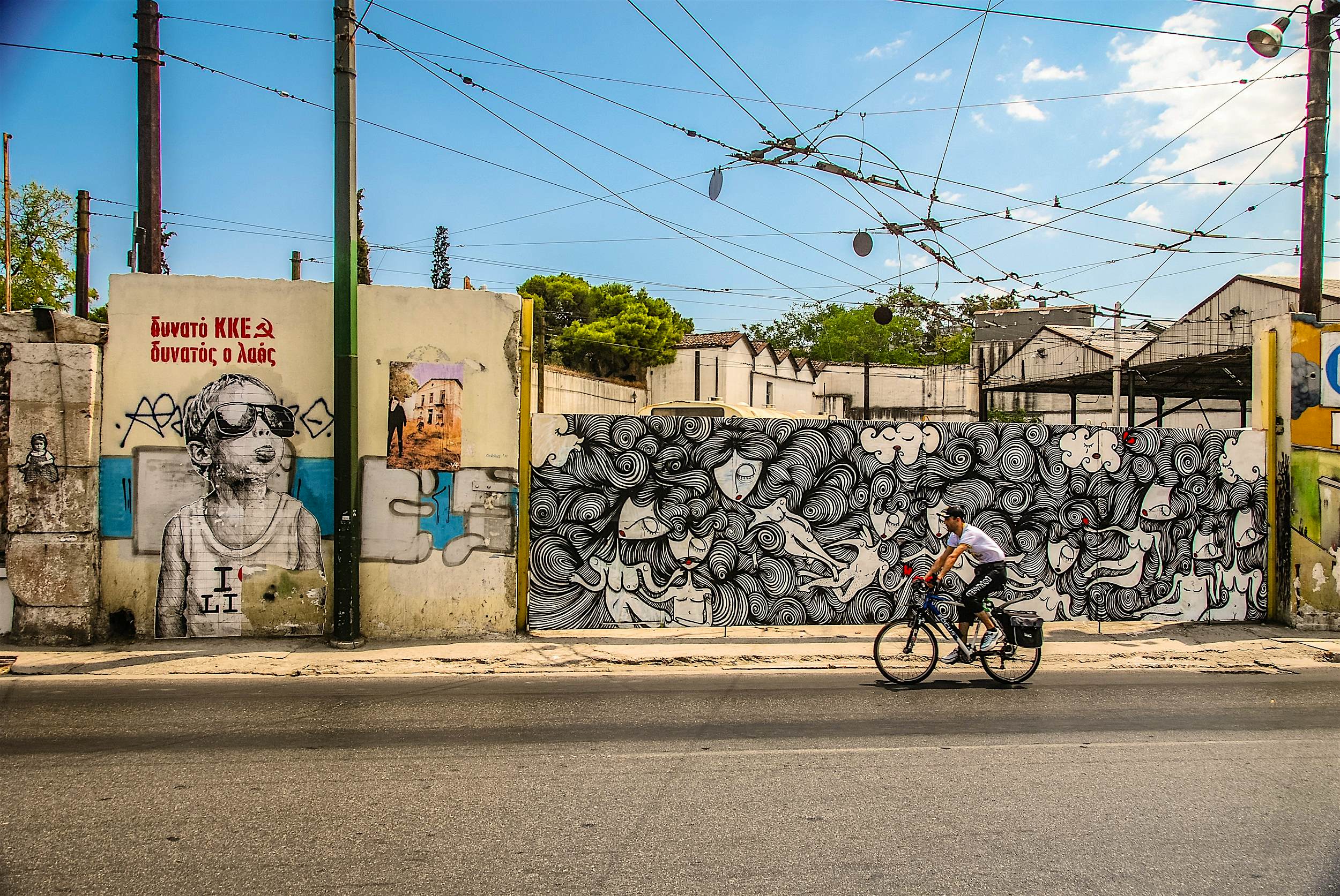 Street art, Athens A cyclist rides past a wall covered in black-and-white illustrations depicting faces and bodies connected by swirls of hair. Another wall has a very large image of a child wearing sunglasses and sticking out their tongue