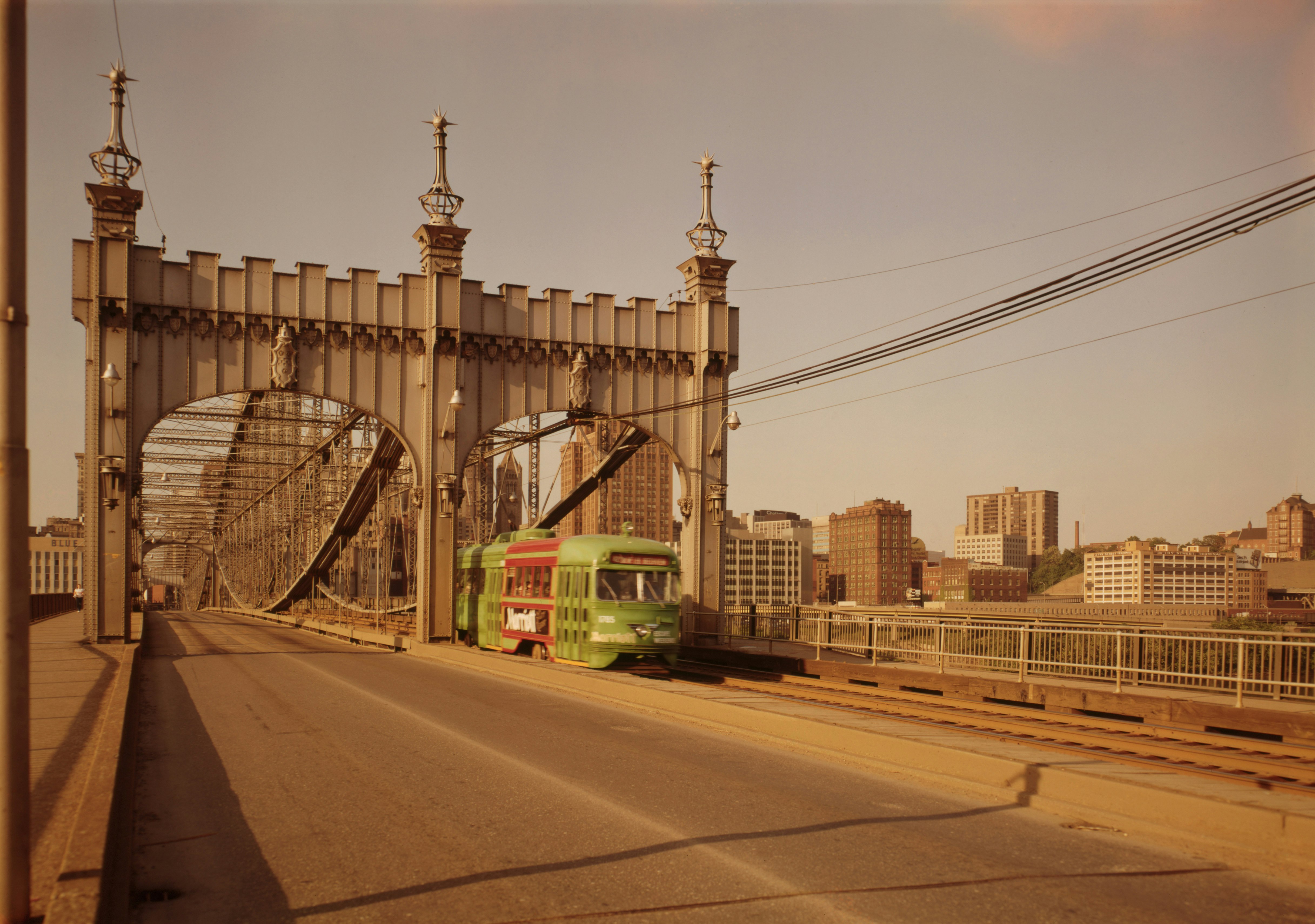 A green, round-faced street car with a red middle section travels across the Smithfield Street Bridge in Pittsburgh in the 1960s. The photo has a warm vintage hue and the bridge is grey steel with ornate crenelations over two arches where the roadway and railway pass.