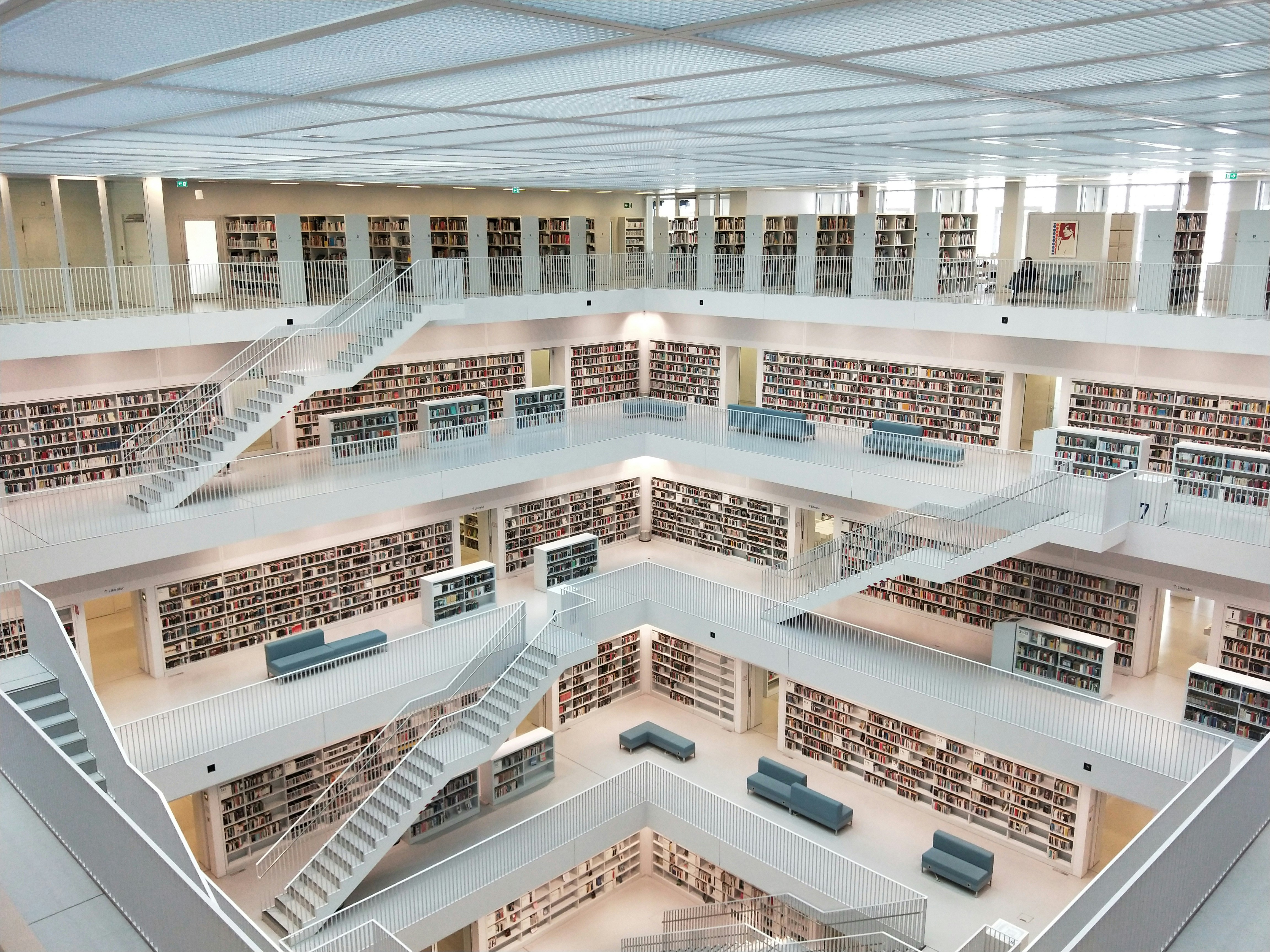 View from an upper gallery down onto a central atrium in a modern public library with glossy white floors, stairs and walls. The walls of each floor are lined floor to ceiling with books, and there are grey sofas and free-standing bookshelves dotted around.