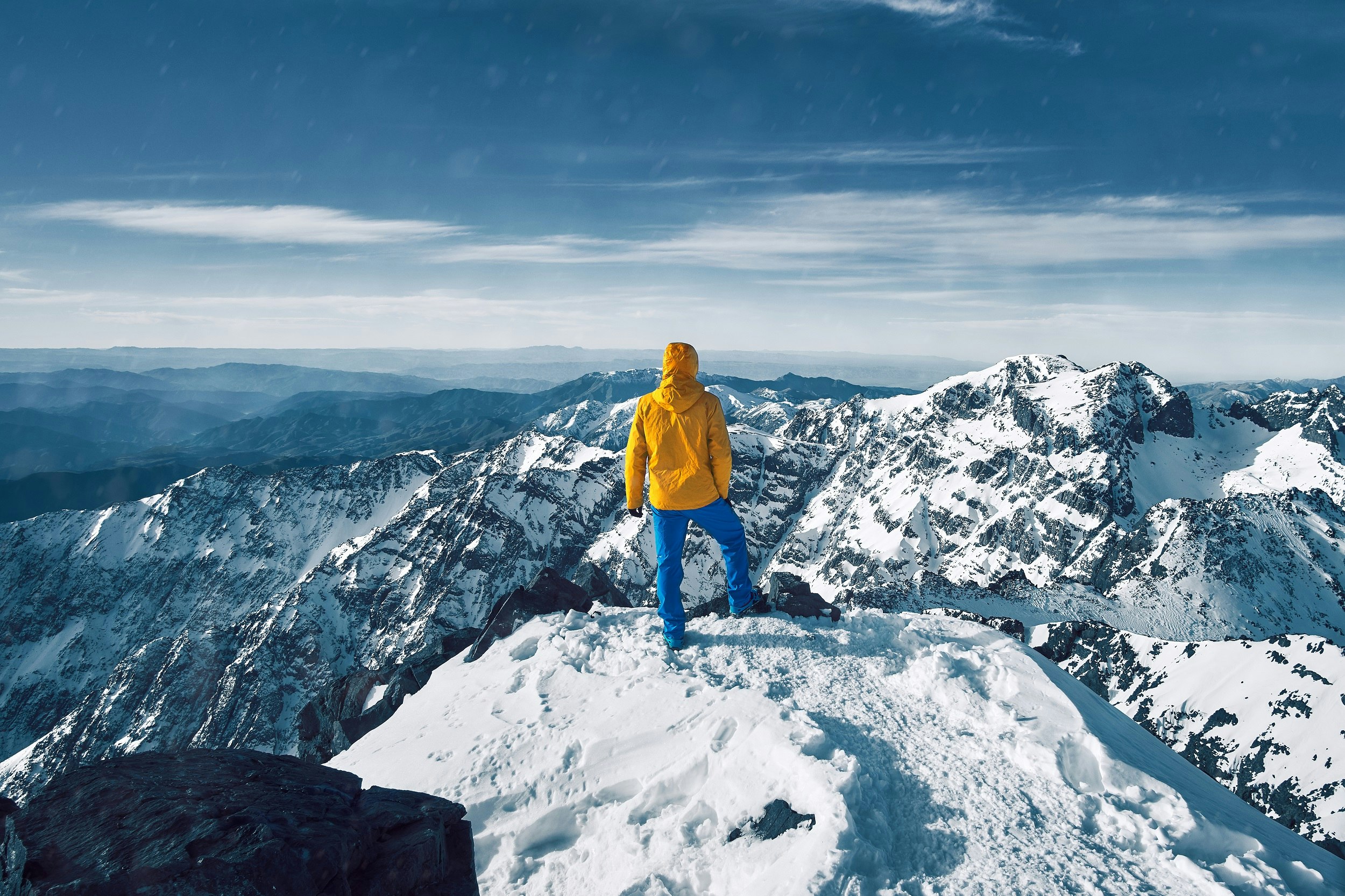 A climber in a yellow jacket and blue trousers stands with their back to the camera on the snowy summit of Toubkal; in the distance are more snow-covered peaks beneath a blue sky.