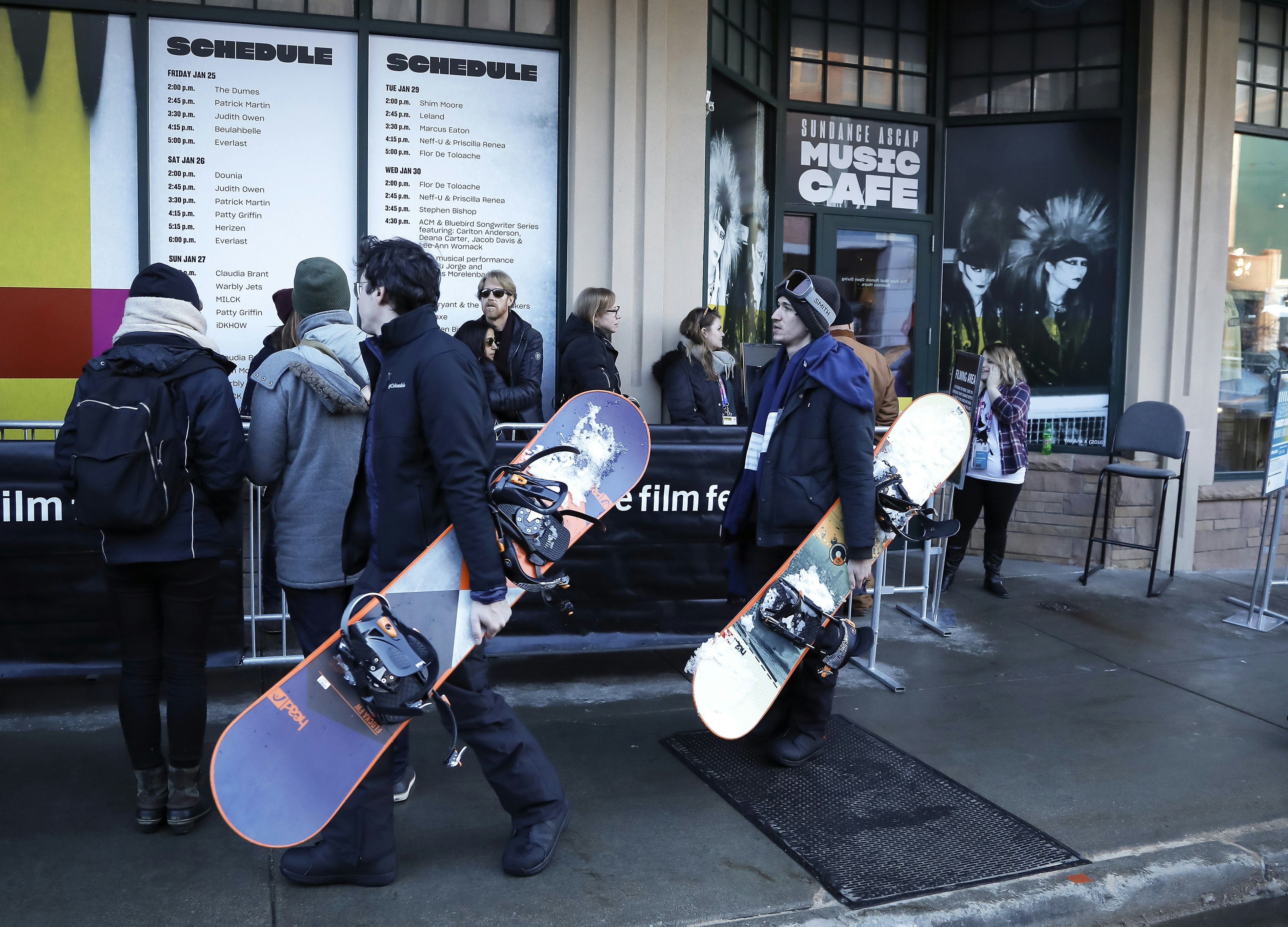 Two men in black ski jackets and snow pants walk down Main Street at the 2019 Sundance Festival with snowboards tucked under their arms. On the other side of a Sundance barrier, film fans wait in line at the Music Cafe