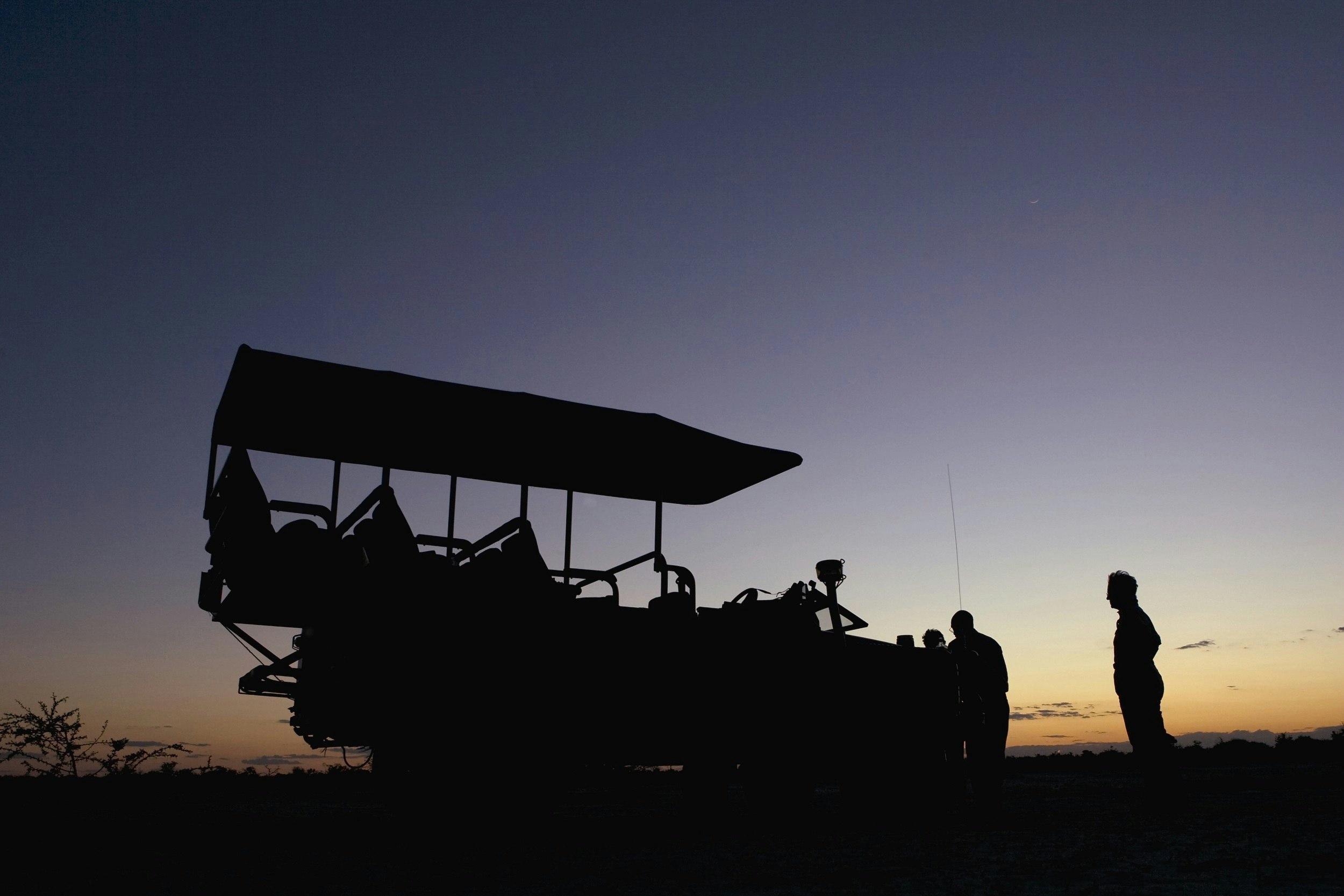 An open-topped 4WD is silhouetted against a purple sky at sunset; a few people stand around the font of it enjoying a drink.