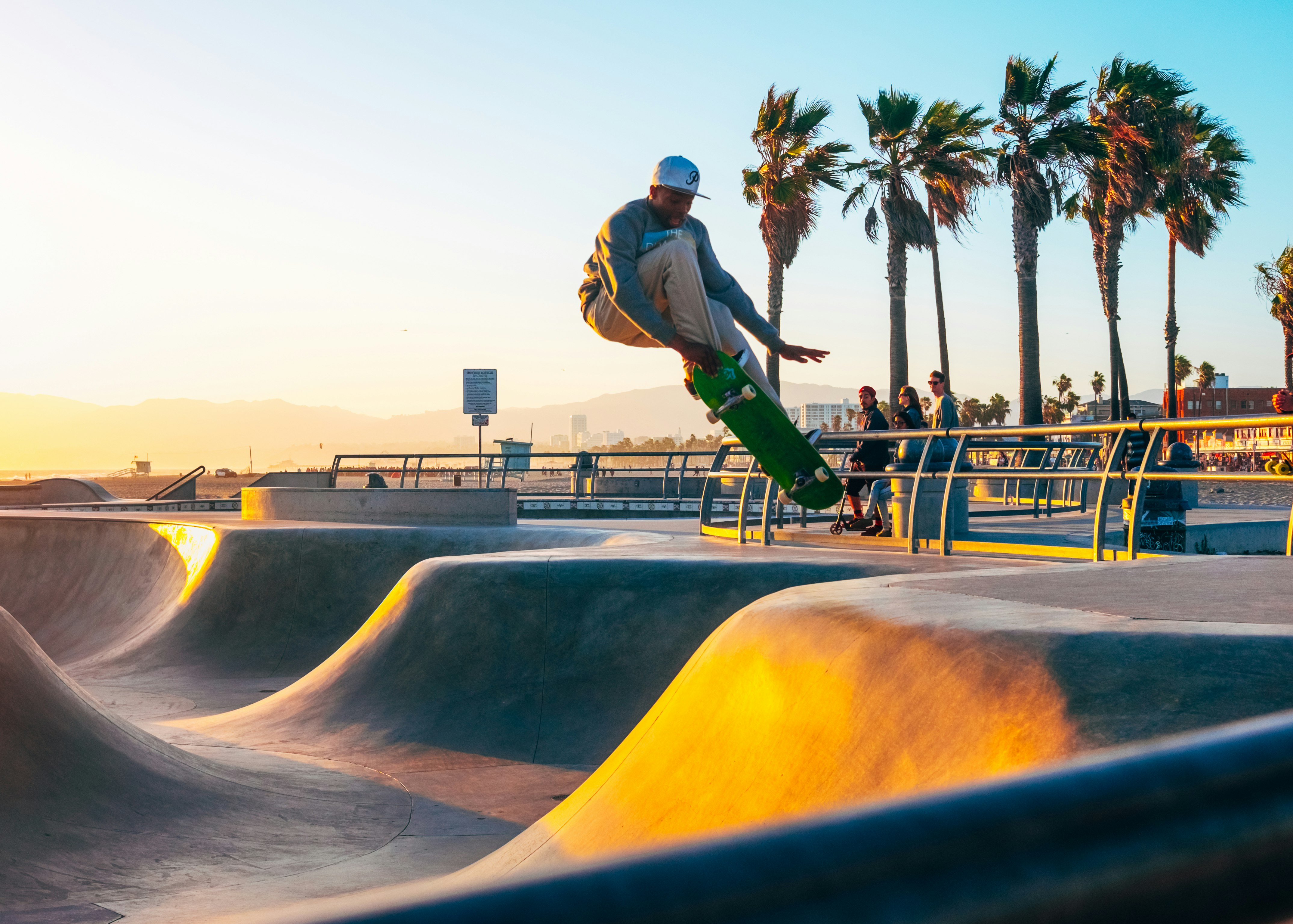 A young man on a skateboard is captured mid-air during a jump at Venice Skate Park; it's sunset and the sun is casting a warm glow on the undulating concrete.