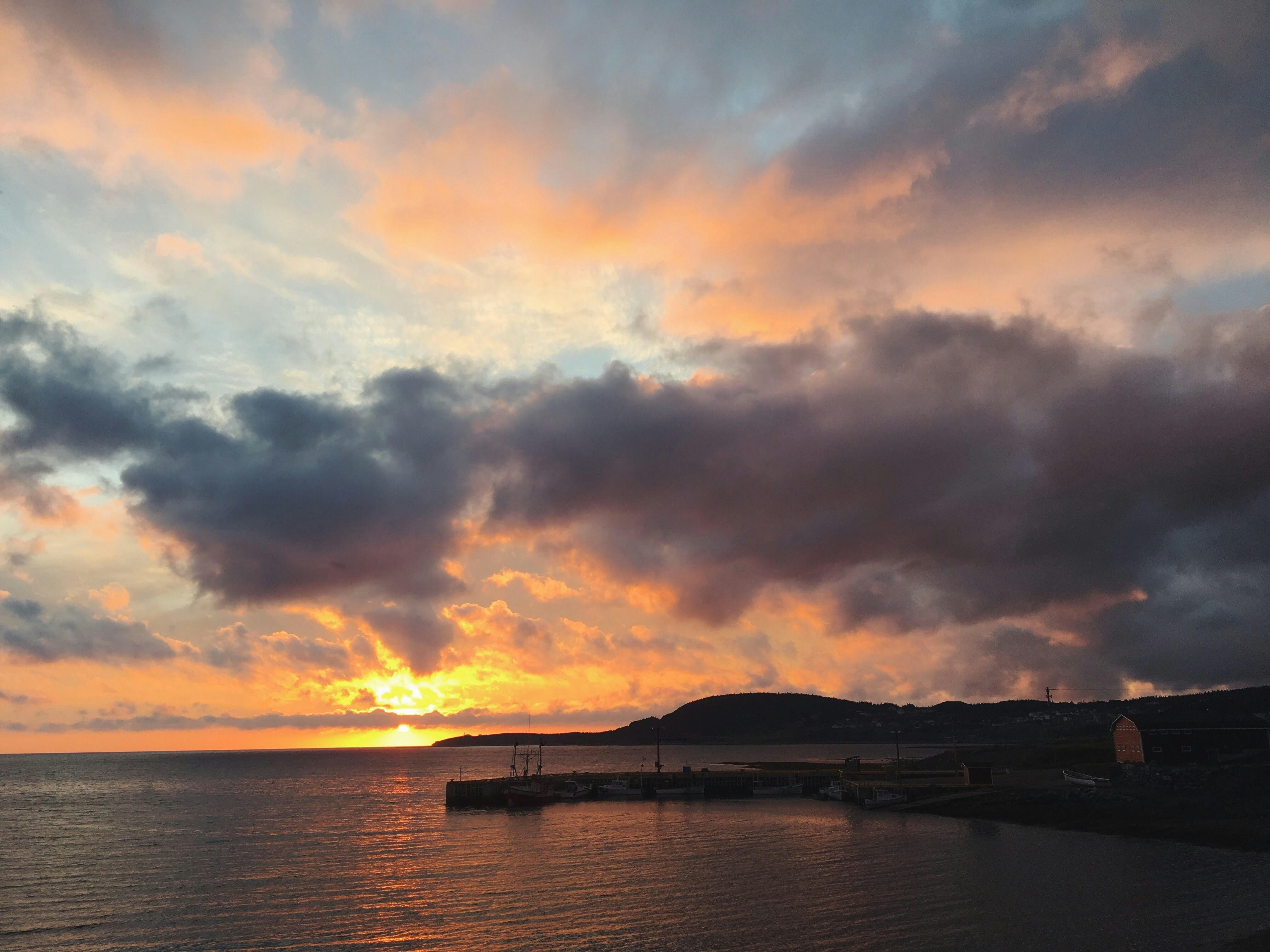 A sunset over a body of water paints a cloudy sky in shades of orange and pink. To the left of the images is a jetty and a simple building.