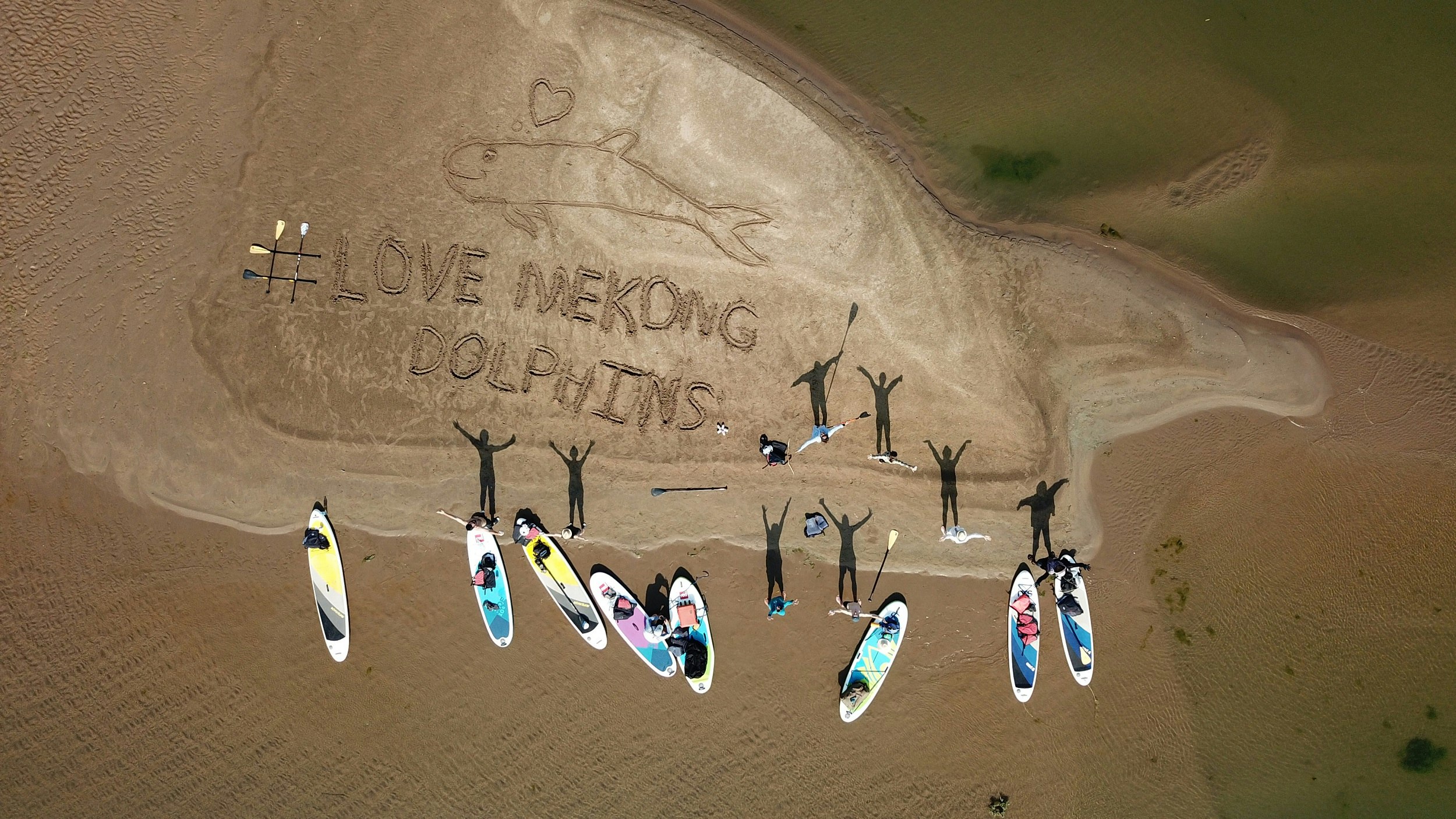A shot looking down at a sandbar in the Mekong River; eight paddle boards sit on the sand and the shadows of the paddlers - with their arms raised - are cast across the scene. In the sand is written "# love mekong dolphins"