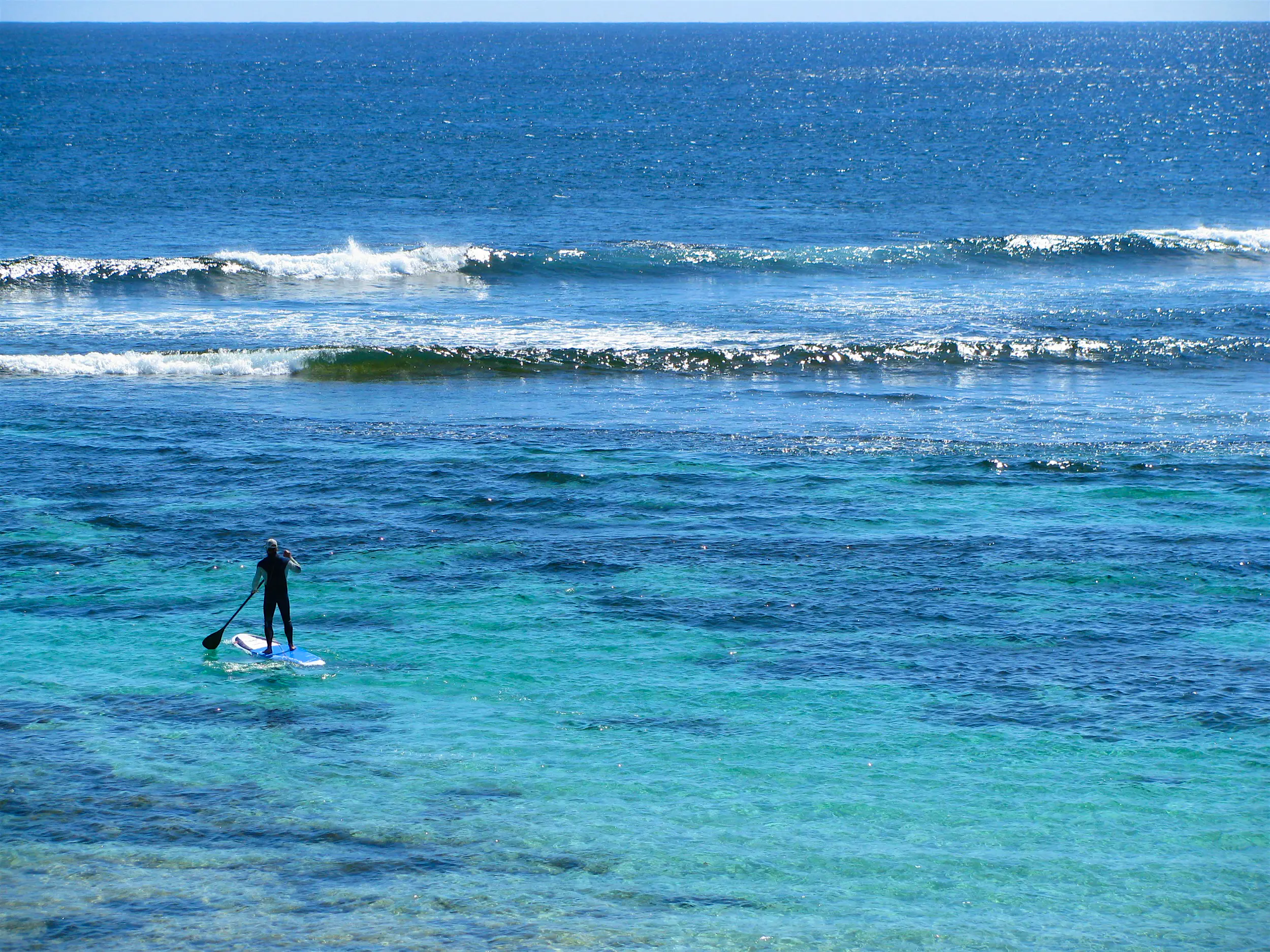 SUP Western Australia A lone person on a stand up paddle board heads through azure shallows towards small crashing waves on the edge of a reef.