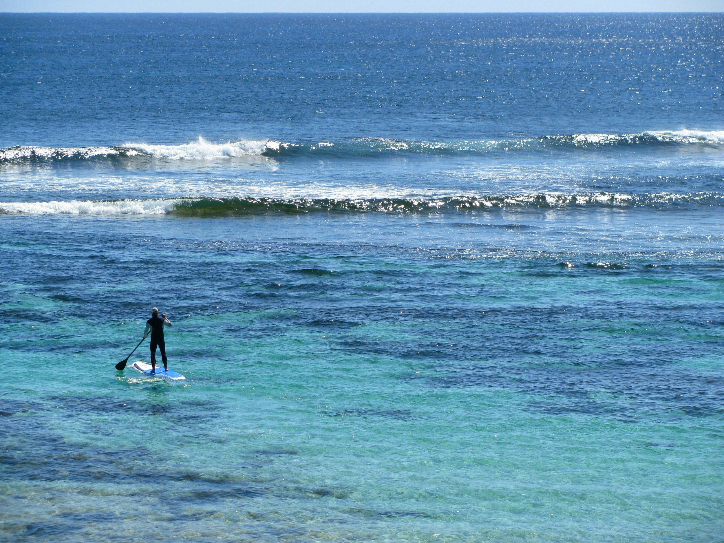 A lone person on a stand up paddle board heads through azure shallows towards small crashing waves on the edge of a reef.