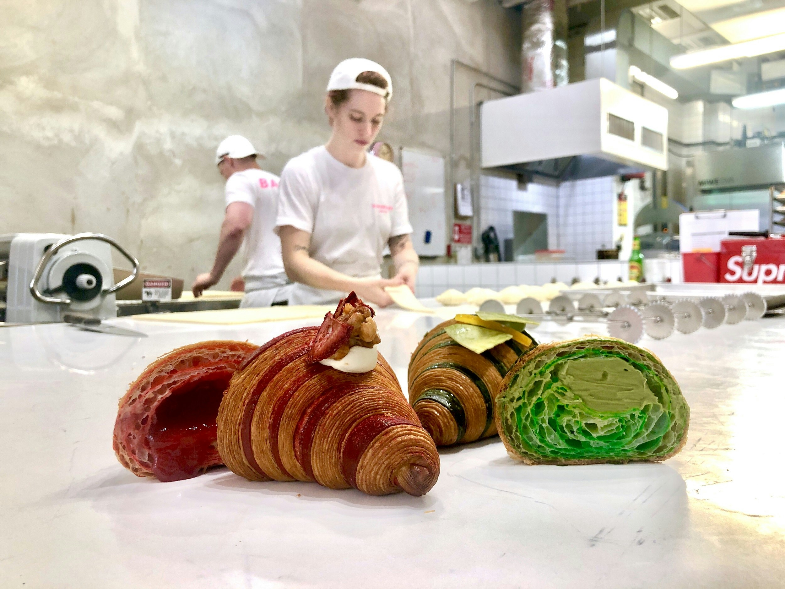 A close-up of two croissants cut in half. One has a red liquid inside, the other is green pastry. Two pastry chefs wearing white t-shirts and hats are working in the background