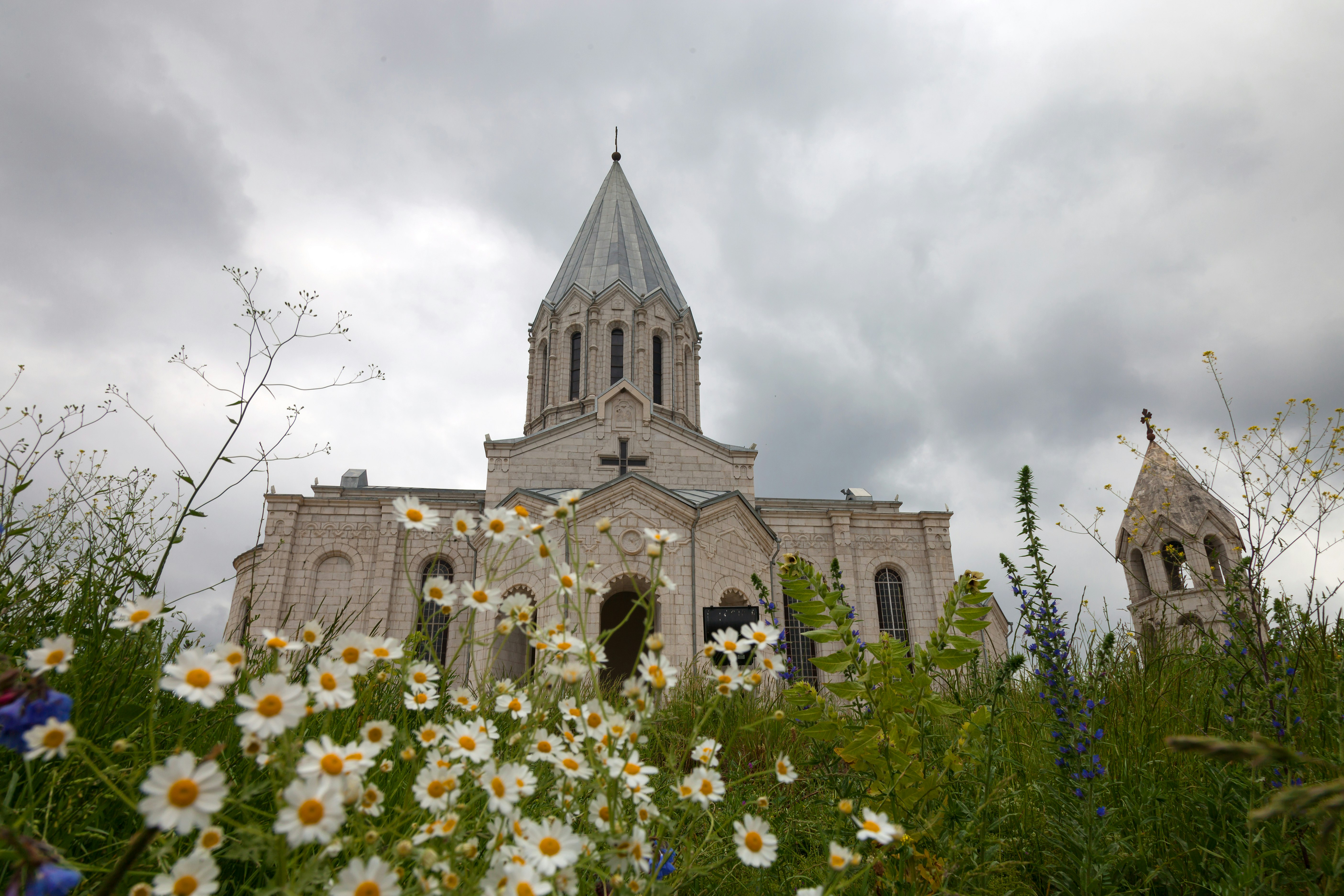 The grey stone exterior of the Ghazanchetsots Cathedral in Shoushi, Artsakh is framed by cloudy skies and a bright patch of chamomile flowers and grass in the immediate foreground 