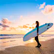 A female surfer standing with a board on the beach at sunset; the sky is cobalt blue at the top of the image, but golden at sea level.
