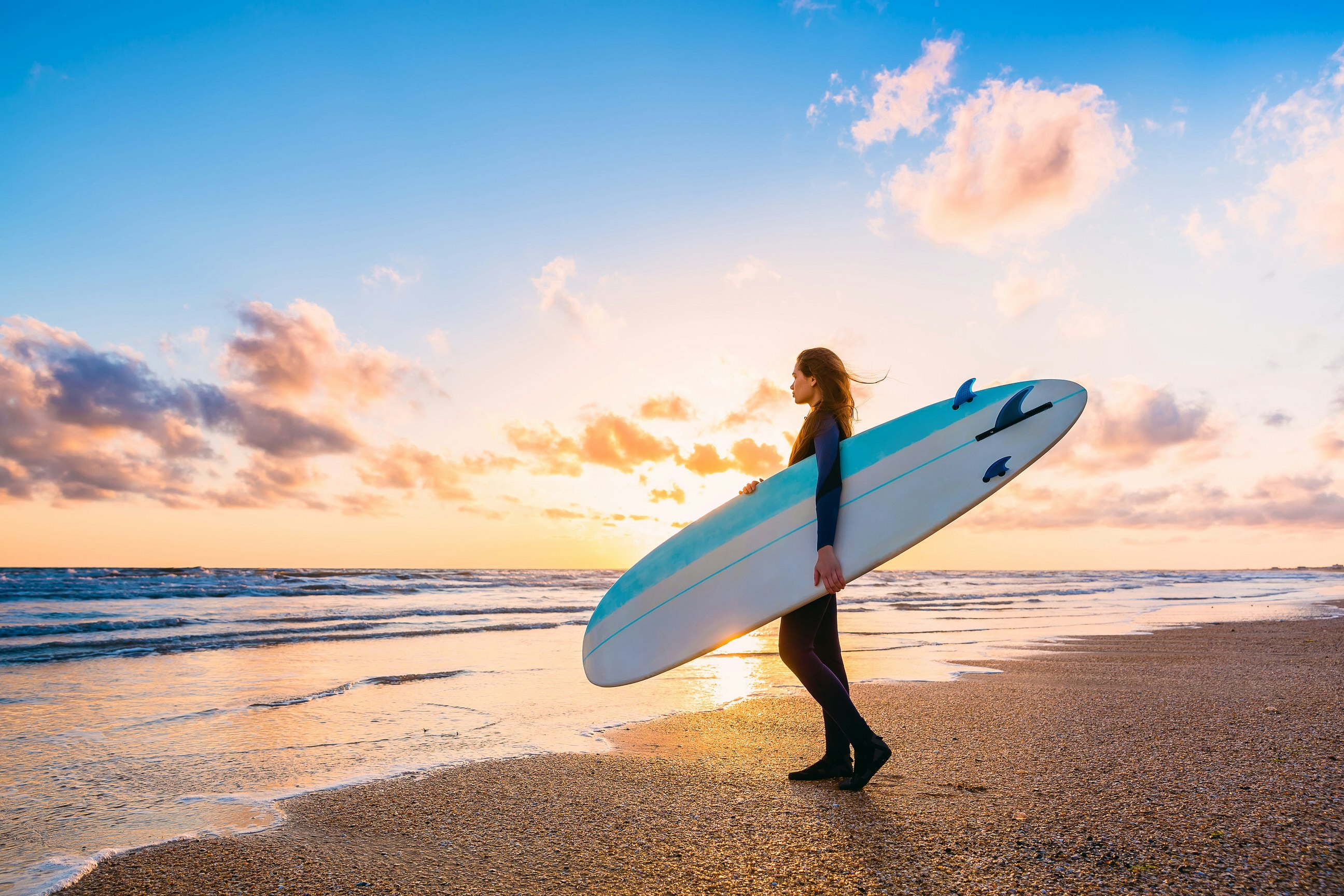 A female surfer standing with a board on the beach at sunset; the sky is cobalt blue at the top of the image, but golden at sea level.