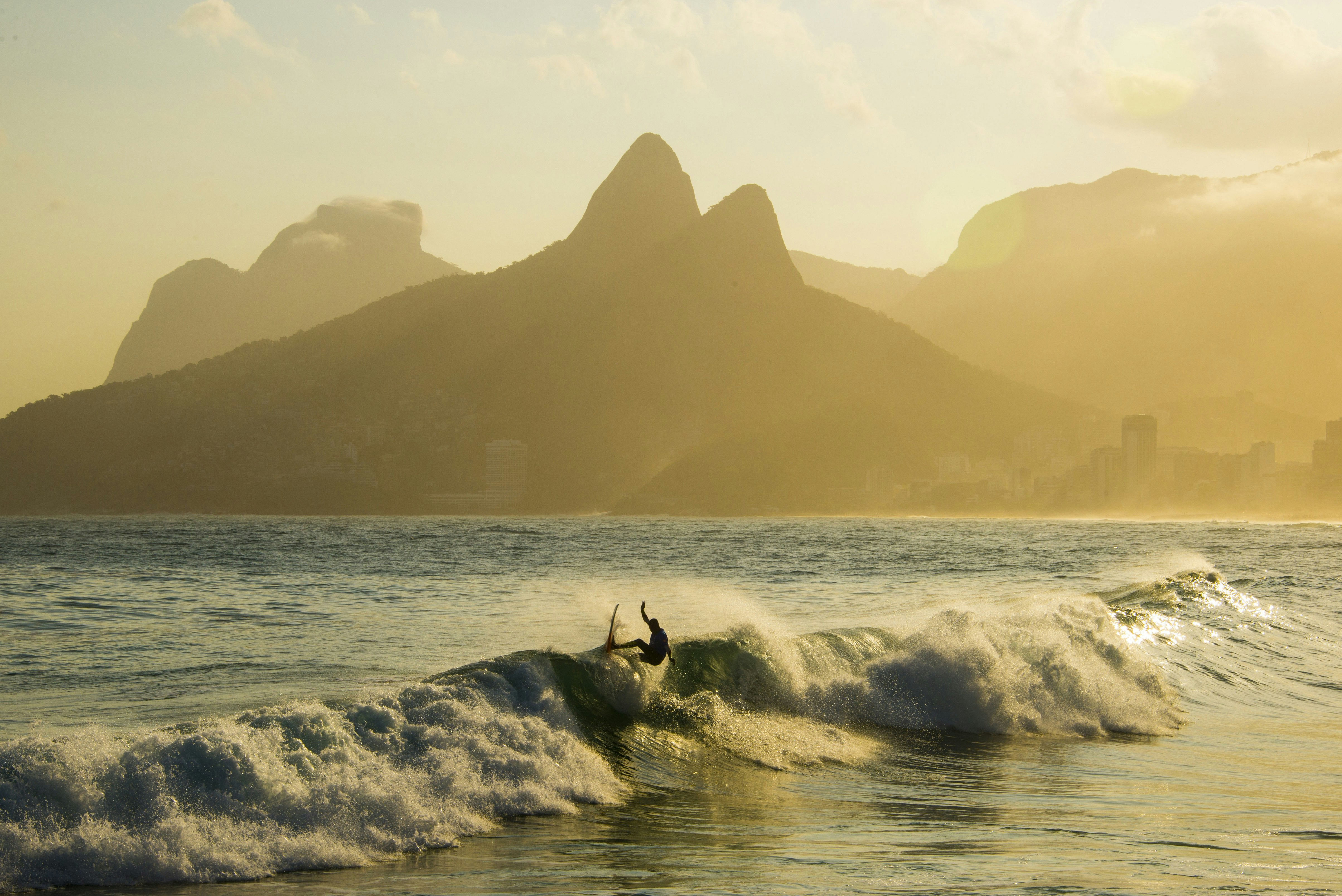 A surfer riding the very top of a crashing wave on Ipanema beach, with the distinctive silhouettes of Rio's mountains in the background.