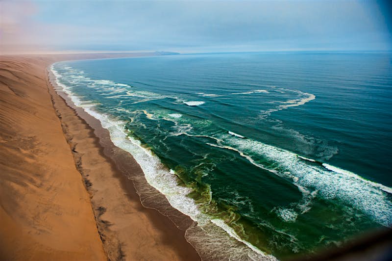 Huge reddish dunes roll down to a beach of the same colour; waves roll in and break atop the greenish blue South Atlantic Ocean.