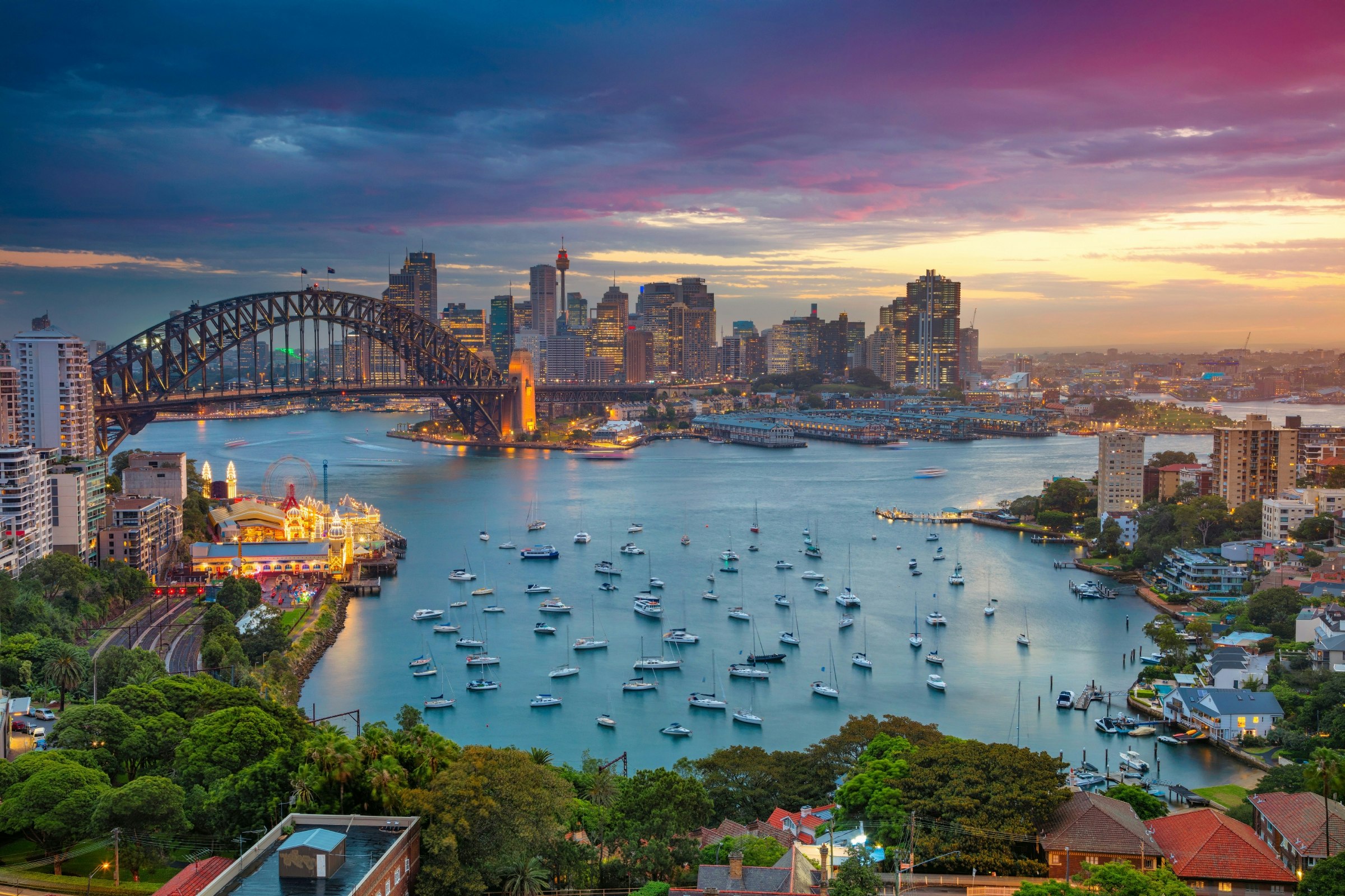 A sunset scene of Sydney Harbour; many boats bob in the water with the iconic Sydney Harbour Bridge in the background.