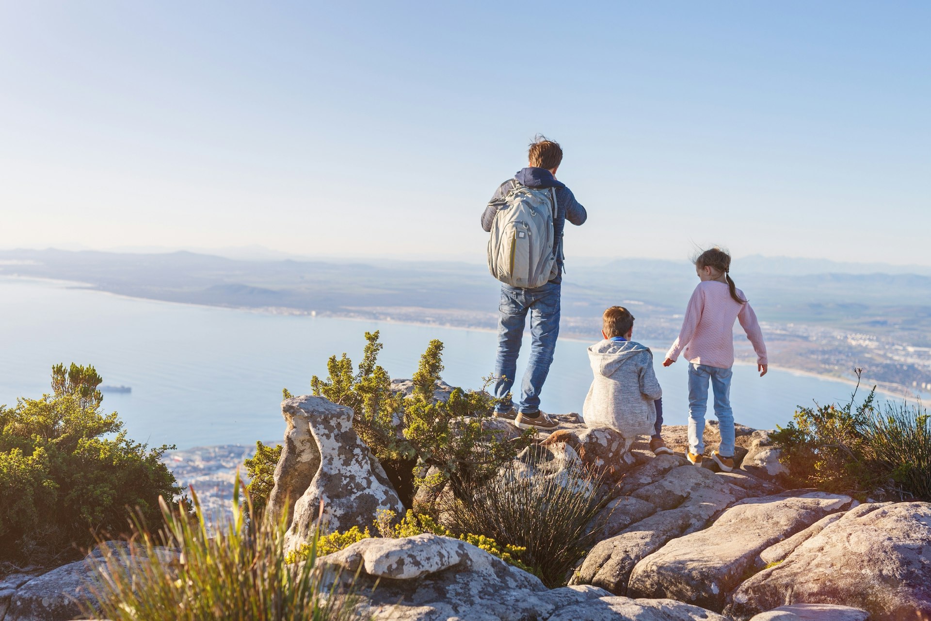 A family with two kids at a rocky lookout over Cape Town on top of Table mountain; below the bay sweeps along to the horizon.