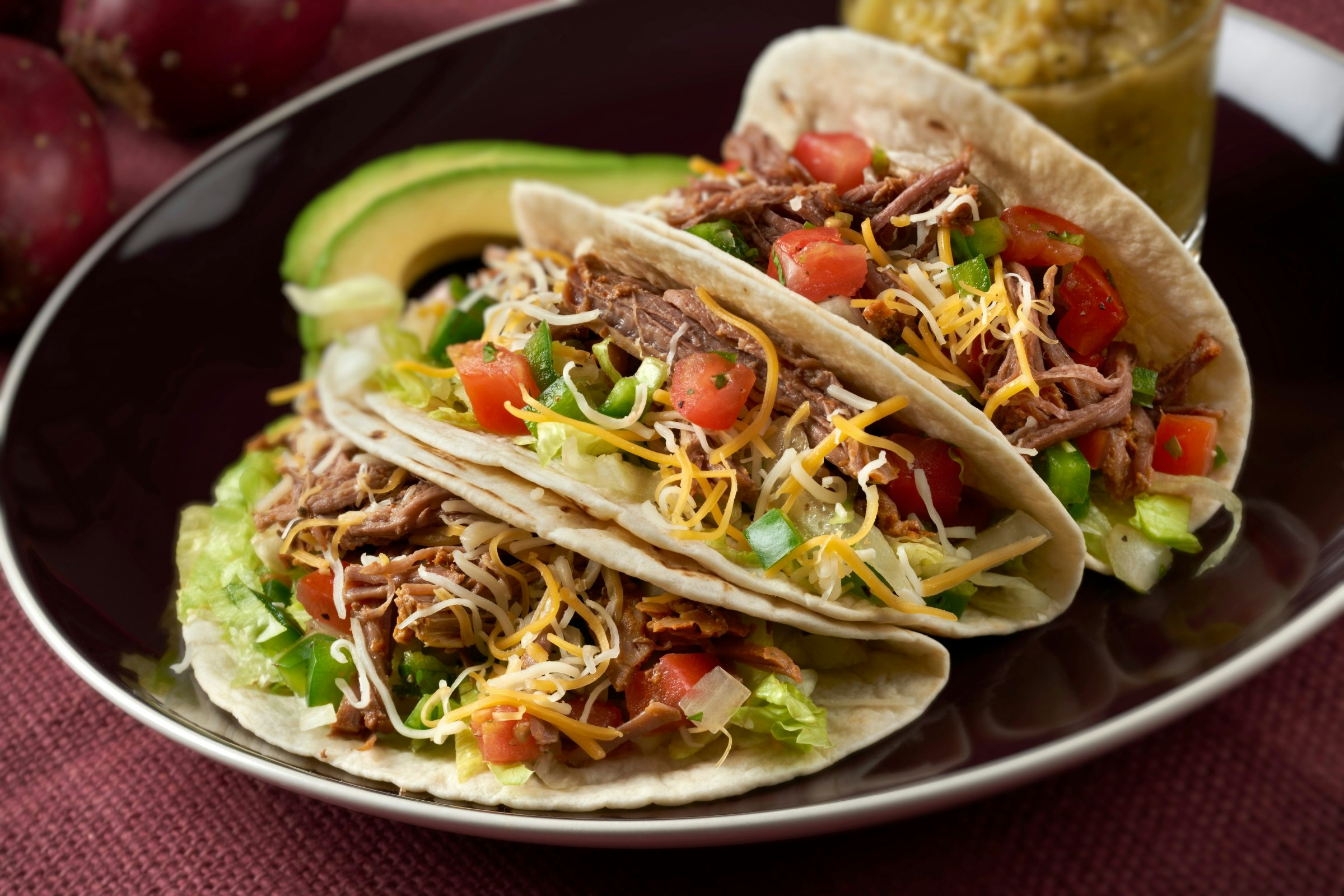 Three meat tacos (with ingredients encased in a corn tortilla) rest on a circular black plate. The tacos are also stuffed with various vegetables and sauces.
