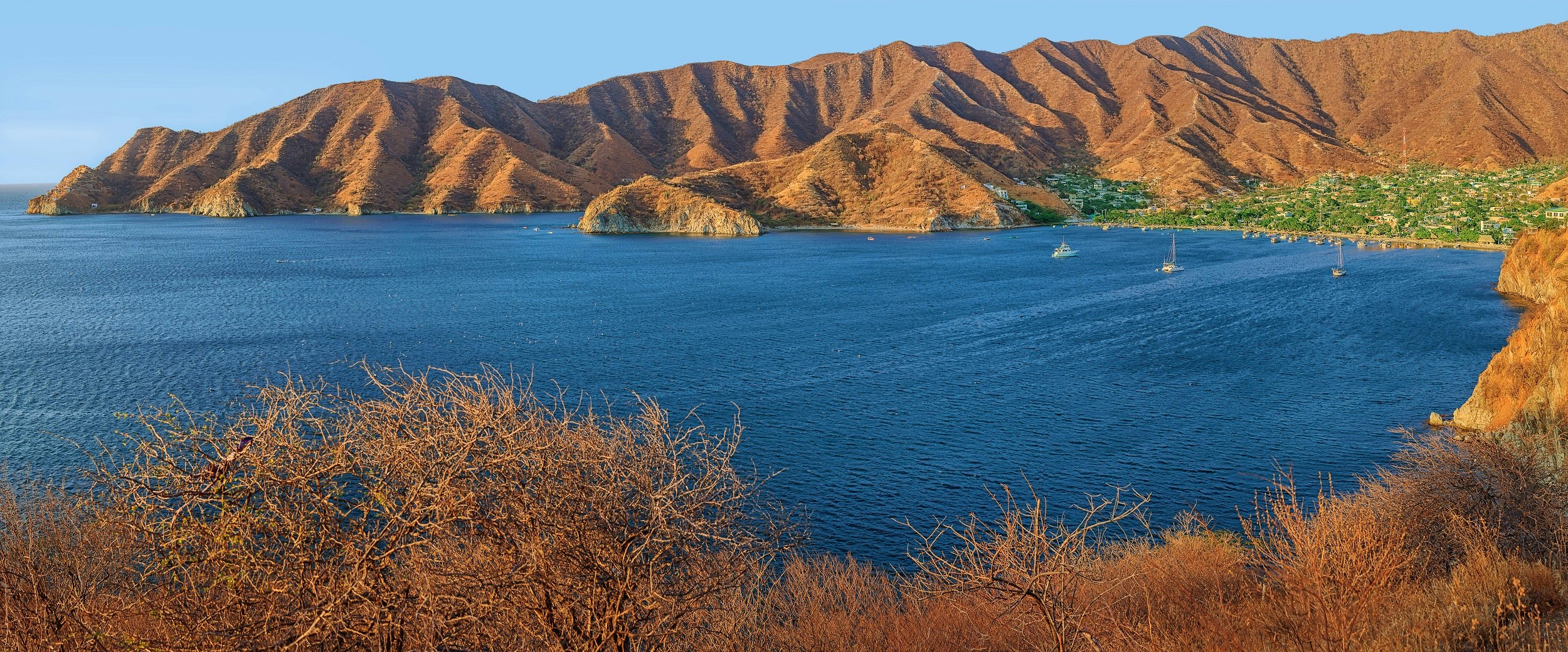 A shot looking down over a large bay; the arms of the bay are made up of orange, almost crenulated hills that plunge to the ocean. The heart of the bay on the shore is a flat patch of green, where the village of Taganga resides.