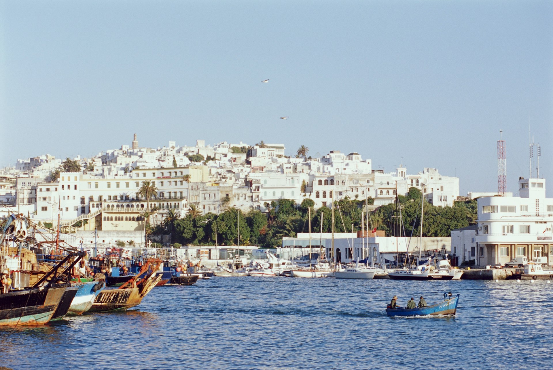 Looking over the waters of the Mediterranean, the white facades of Tangier's old city rises from the shoreline; boats are moored to shore, while a small one makes its way across the harbour.