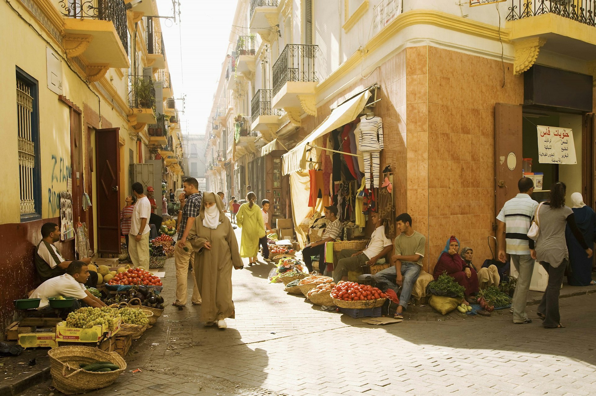 A shot looking down a crossroads of some intersecting alleys in Tangier; the light is warm and there are many people milling about outside shops.