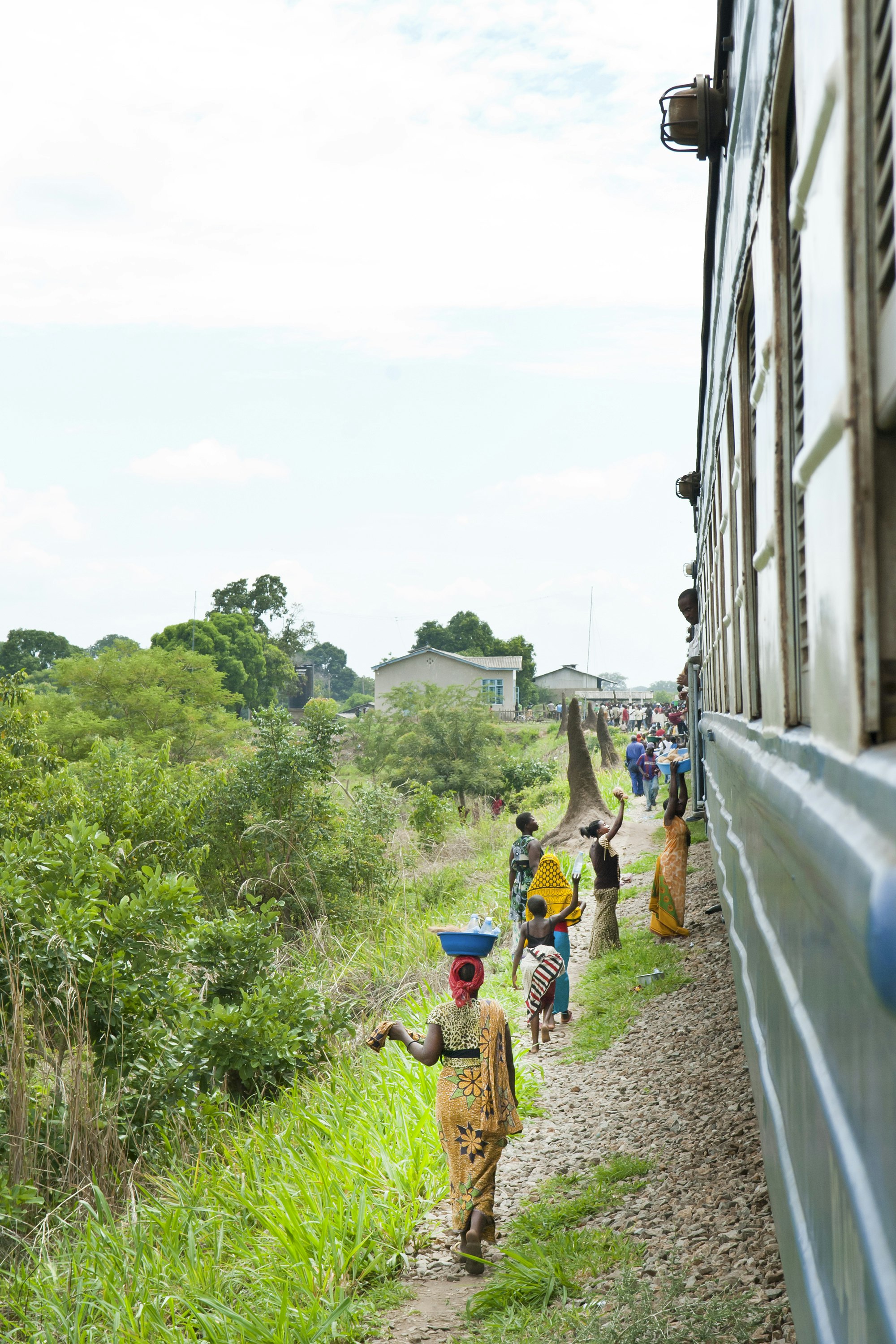 Women walk along the gravel verge beside a stopped train; they carry bowls on their heads and sell drinks and food through the windows of the train.