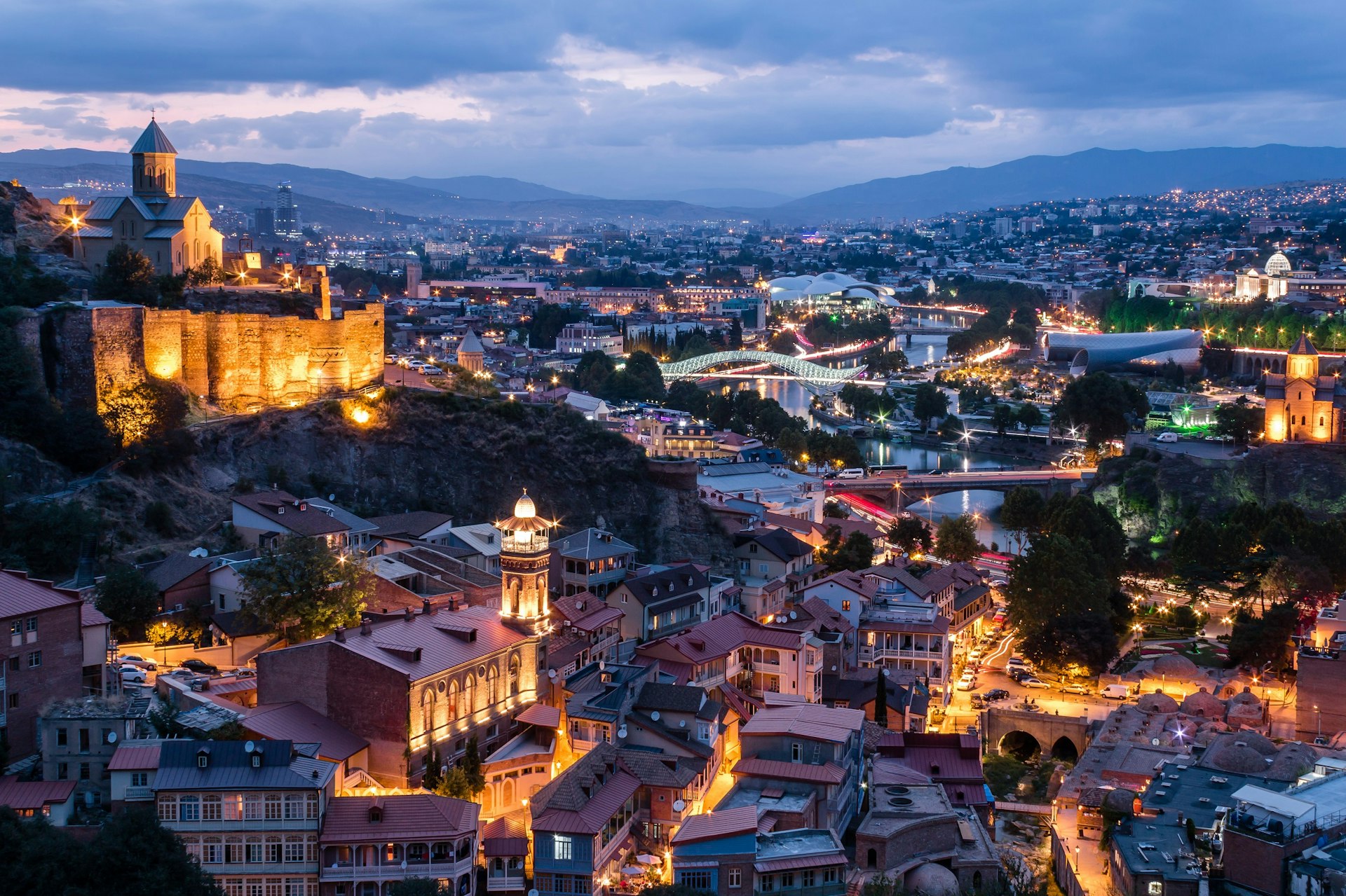 City view at dusk. A hilltop church with fortress-like walls is lit up to the left. There is a river with two illuminated bridges.