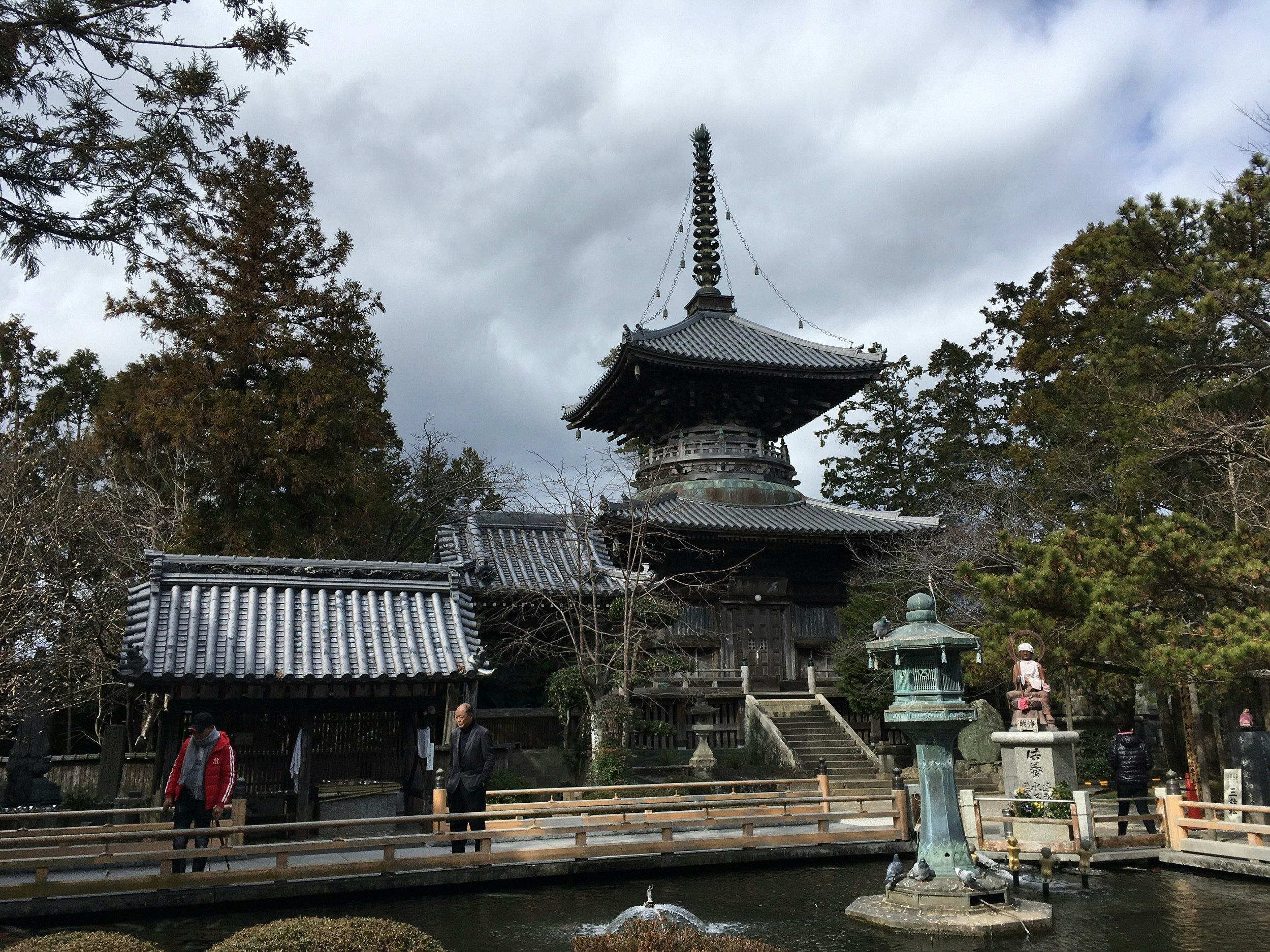 A pagoda building with slate-grey roofs. Two people stand in the foreground looking in a pond.