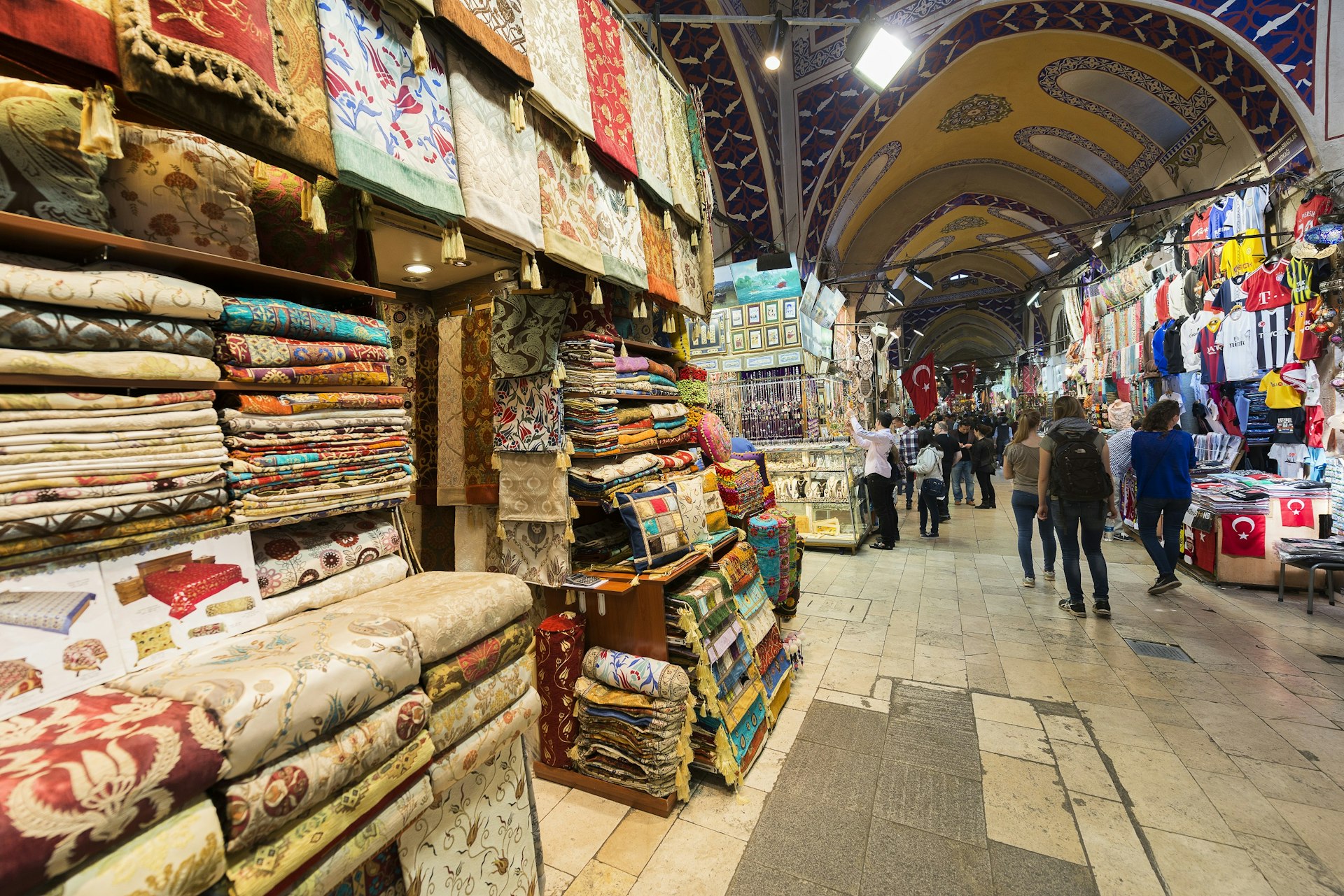 A display outside a shop within a large covered market shows many different heavily patterned textiles for sale