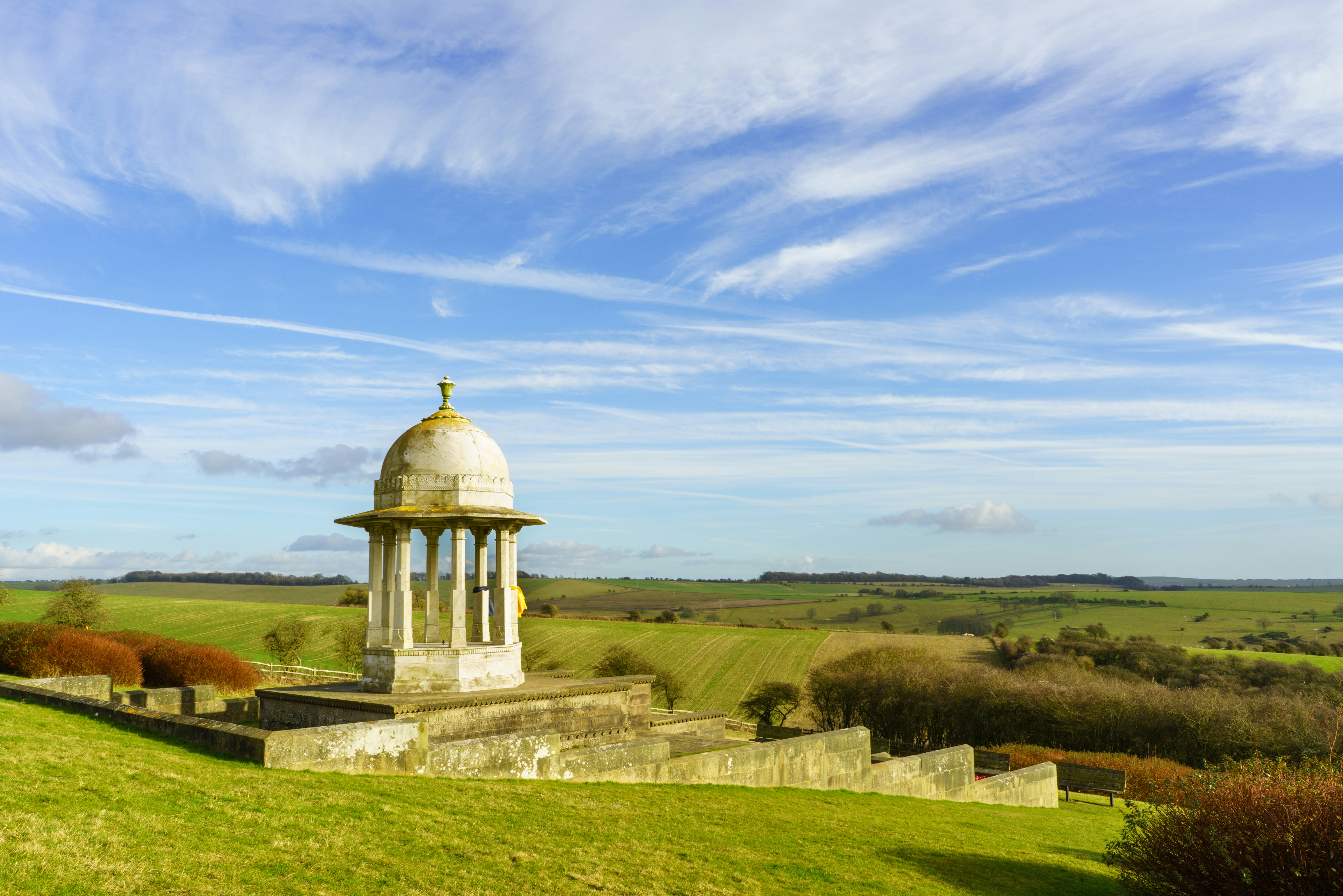 A domed pagoda made from white marble stands amid rolling green hills. The blue sky is criss-crossed with wisps of white cloud.