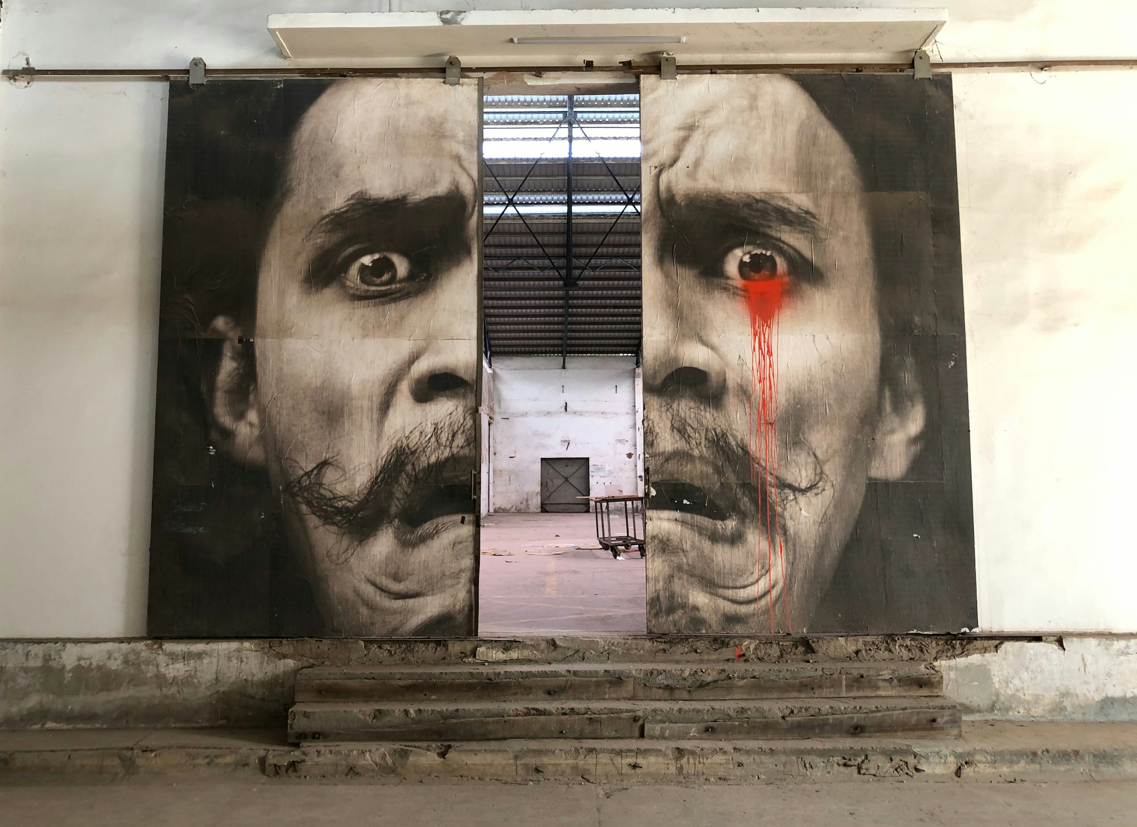 A mural of Salvador Dalí has been painted on two huge doors on rollers; the doors are partly open, so his contorted face is pulled apart. Through the doors is an almost empty, open industrial space.