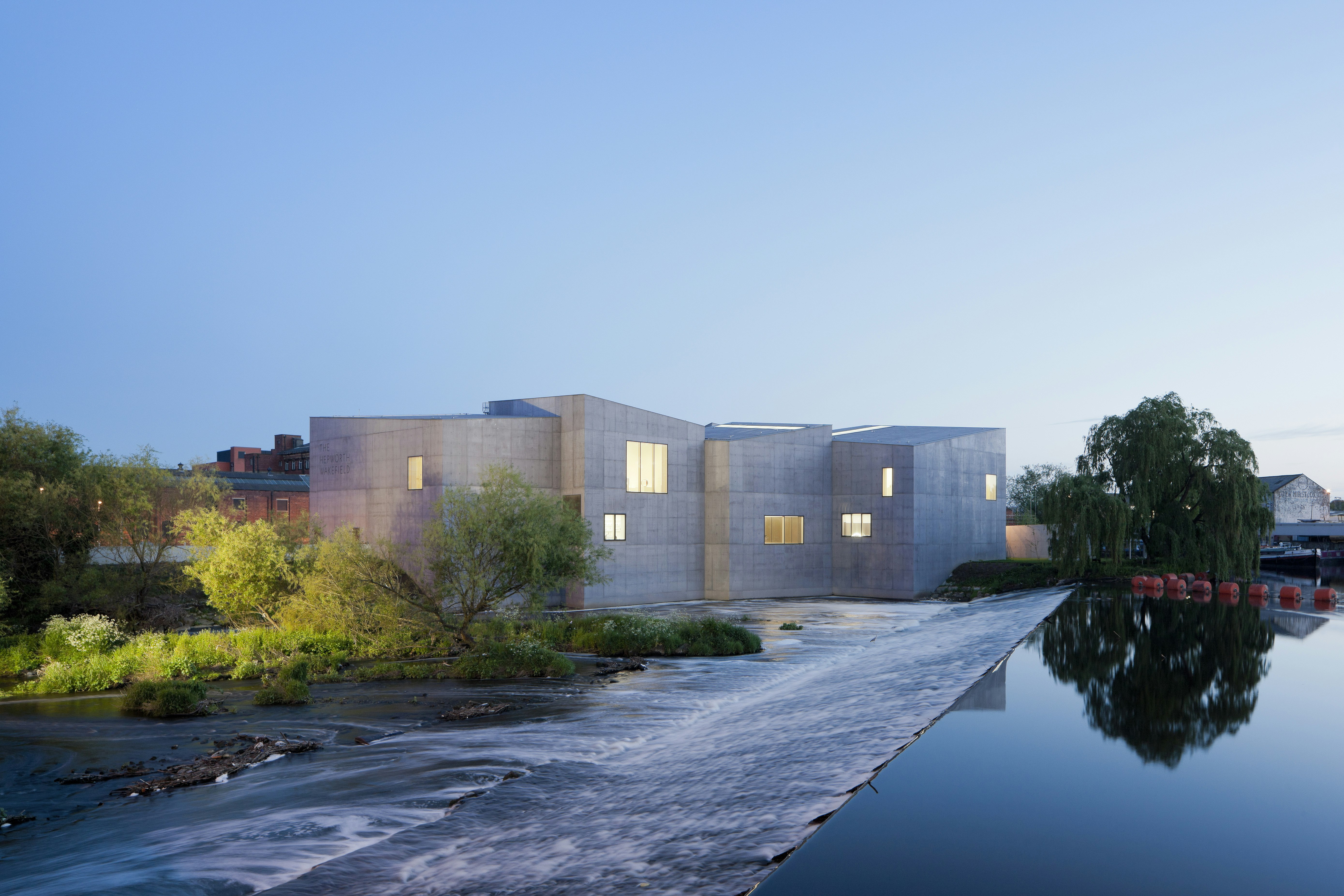 A shot of the minimalist, block-like concrete structure that is The Hepworth Wakefield; trees soften the stark exterior and a glass-like river tumbles down a gentle slope alongside the gallery.