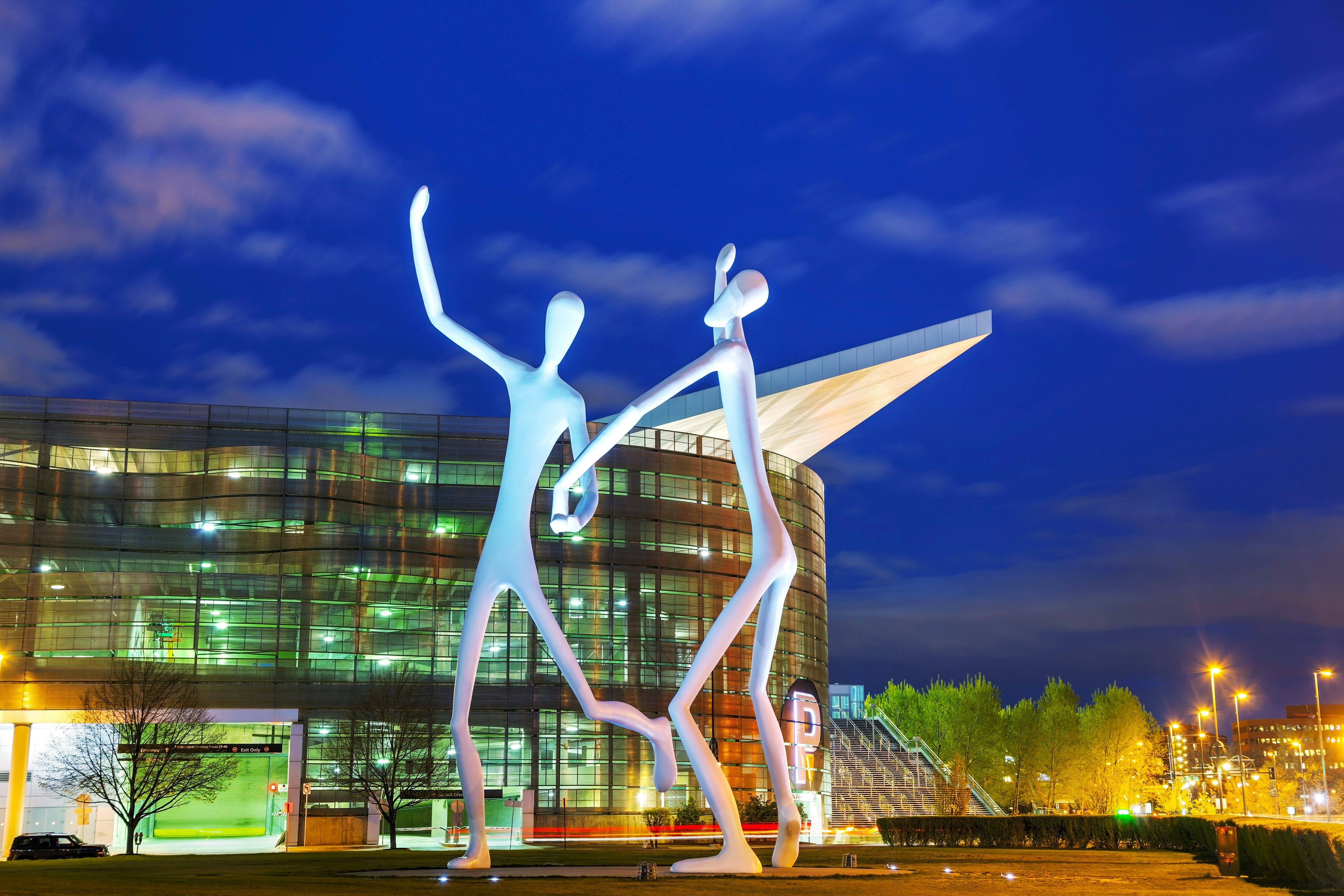 Tall human-like sculptures stand in front of a glass building under a night sky