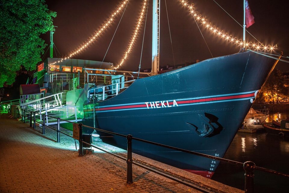 The exterior of the boat Thekla in Bristol; festoon lights are strung from its bow to the main deck.