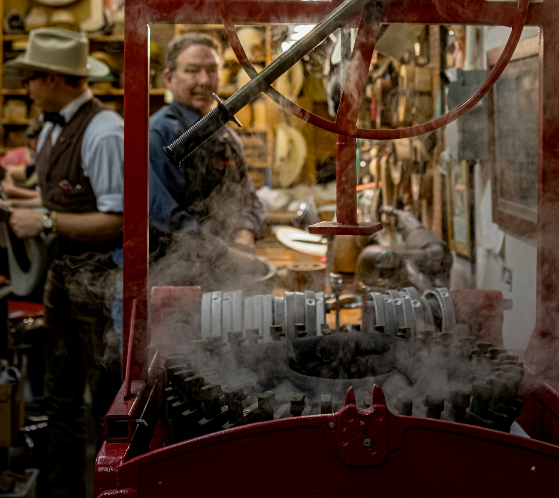 Steam rises around a piece of hat-making equipment with a bright red frame. In the background, Jack Kellogg and Austin Kitchen work on other hats and can be seen just beyond the silver metal components of the old finger blocker machine, which look a little like piano keys.