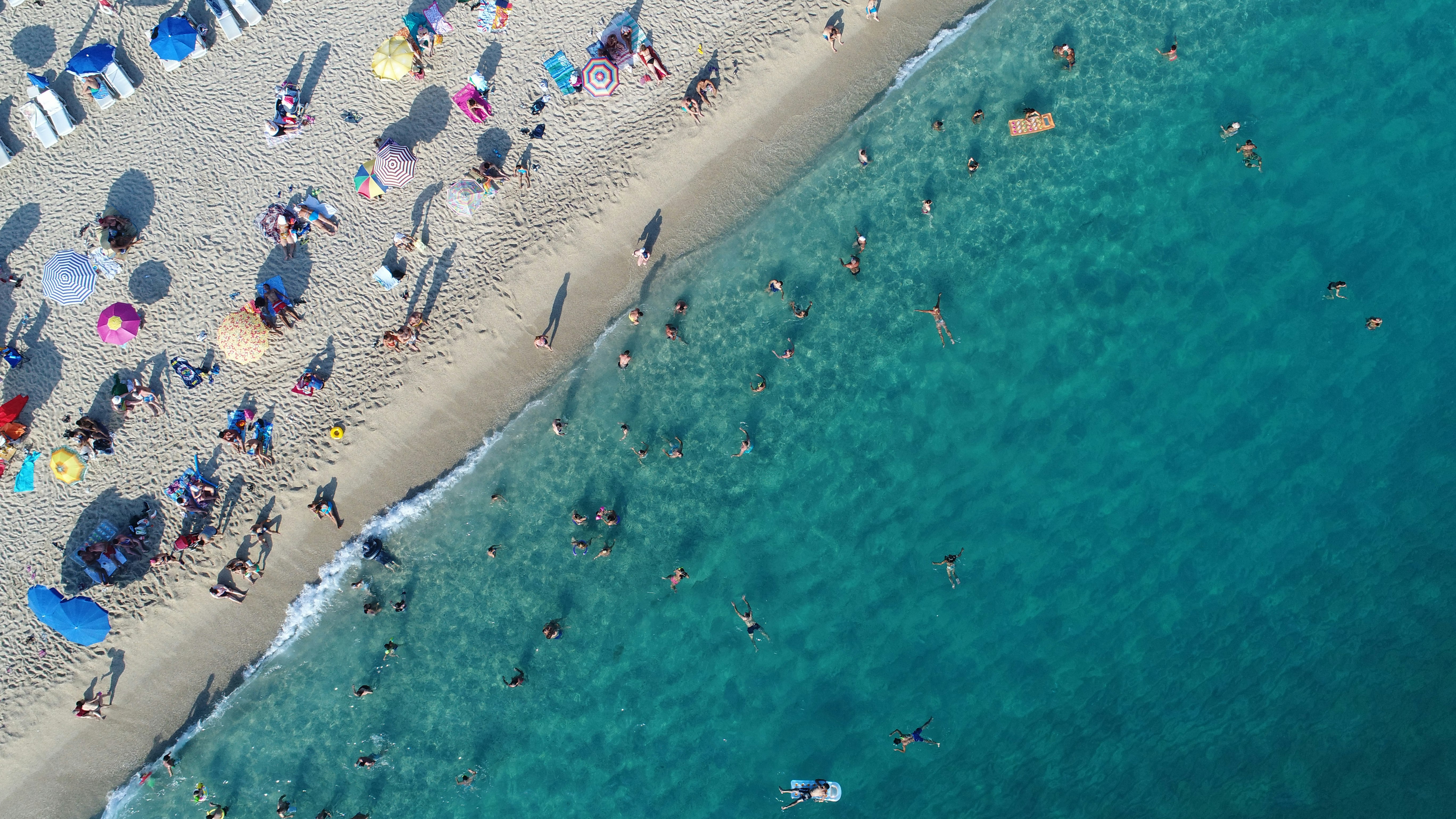 An aerial view of a beach shows colourful umbrellas on the shore and swimmers in the water. 