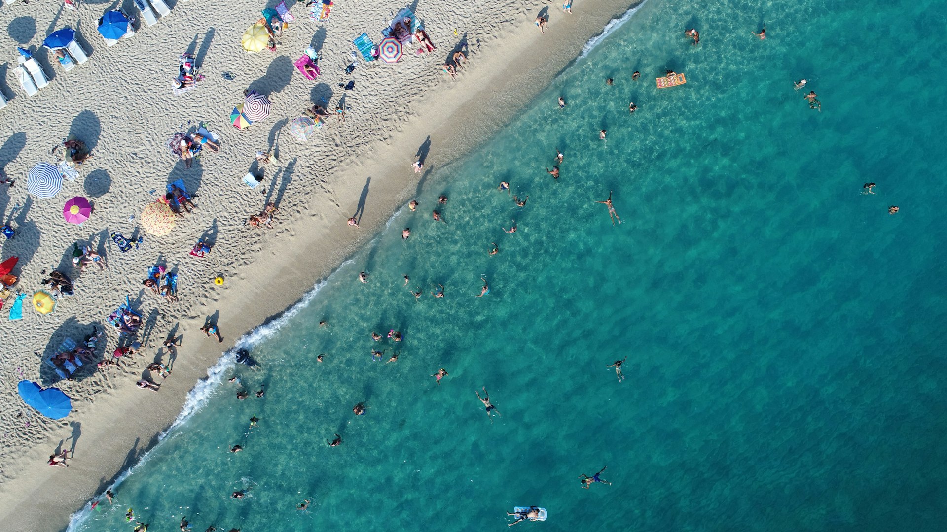 An aerial view of a beach shows colourful umbrellas on the shore and swimmers in the water. 
