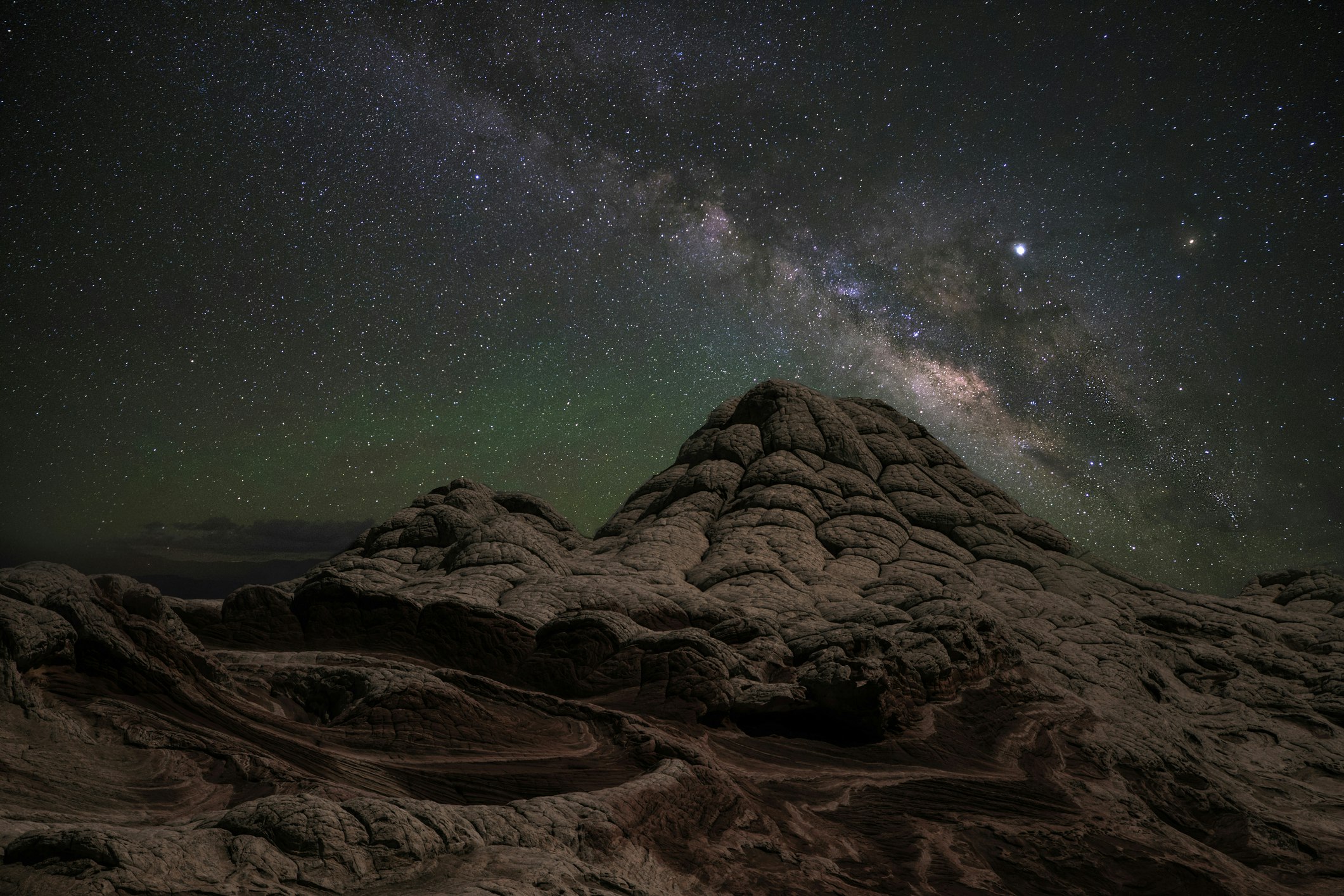 A night sky in shades of blue, green, and purple with a long streak of the Milky Way extending from the top center of the frame to the bottom right hangs over a large sandstone outcropping with whorls and bubbly lumps that form two conical mounds in the center of the shot
