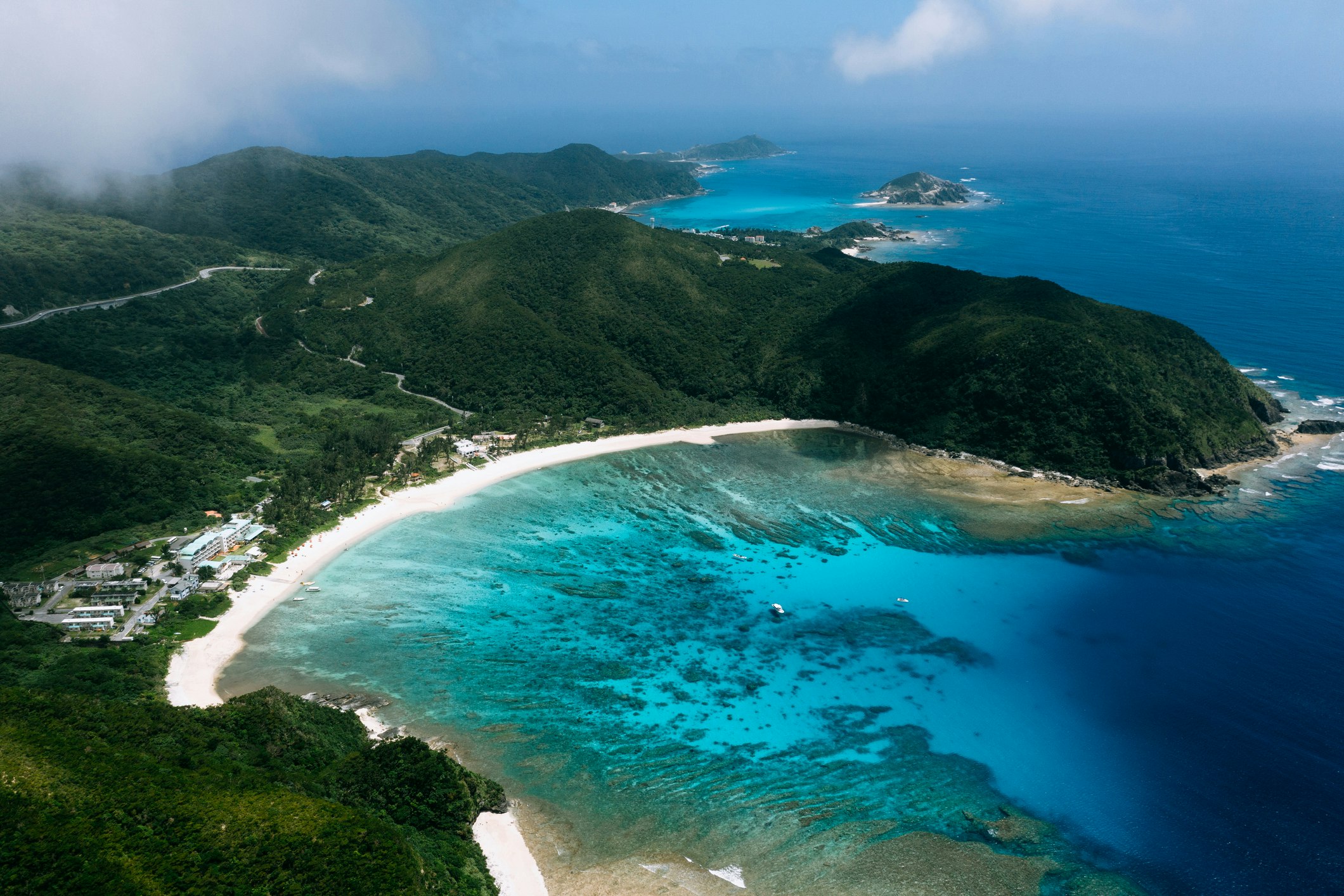 The brilliant blue and teal waters of Keramashoto National Park extend out from a crescent shaped beach, with a thin strip of white sand pressed against the green and black hills of Tokashiki island 
