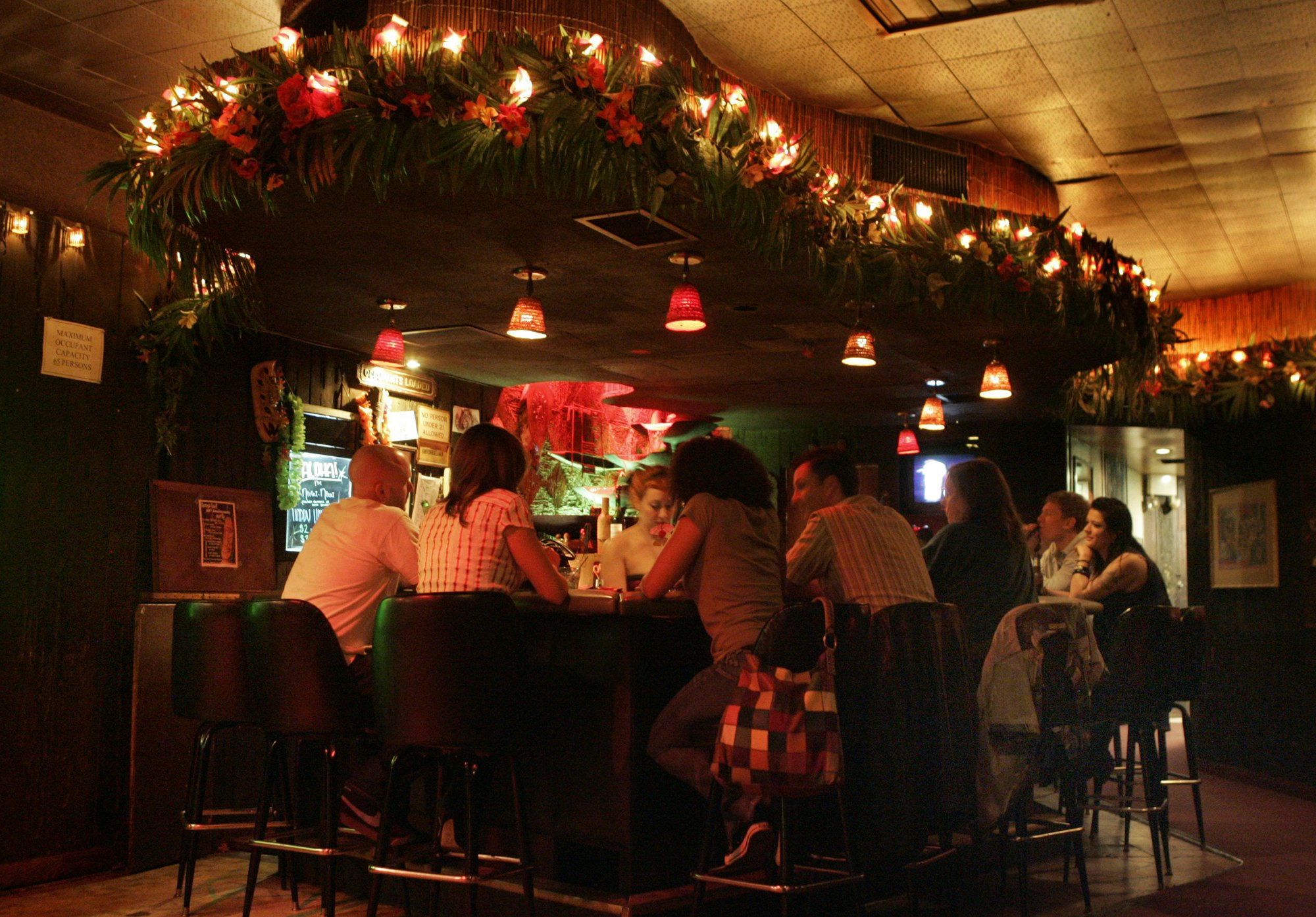 Six patrons sit around the bar at Tonga Hut in Los Angeles, where red tropical flowers are intertwined in a palm frond garland studded with lights over mid-century style barstools