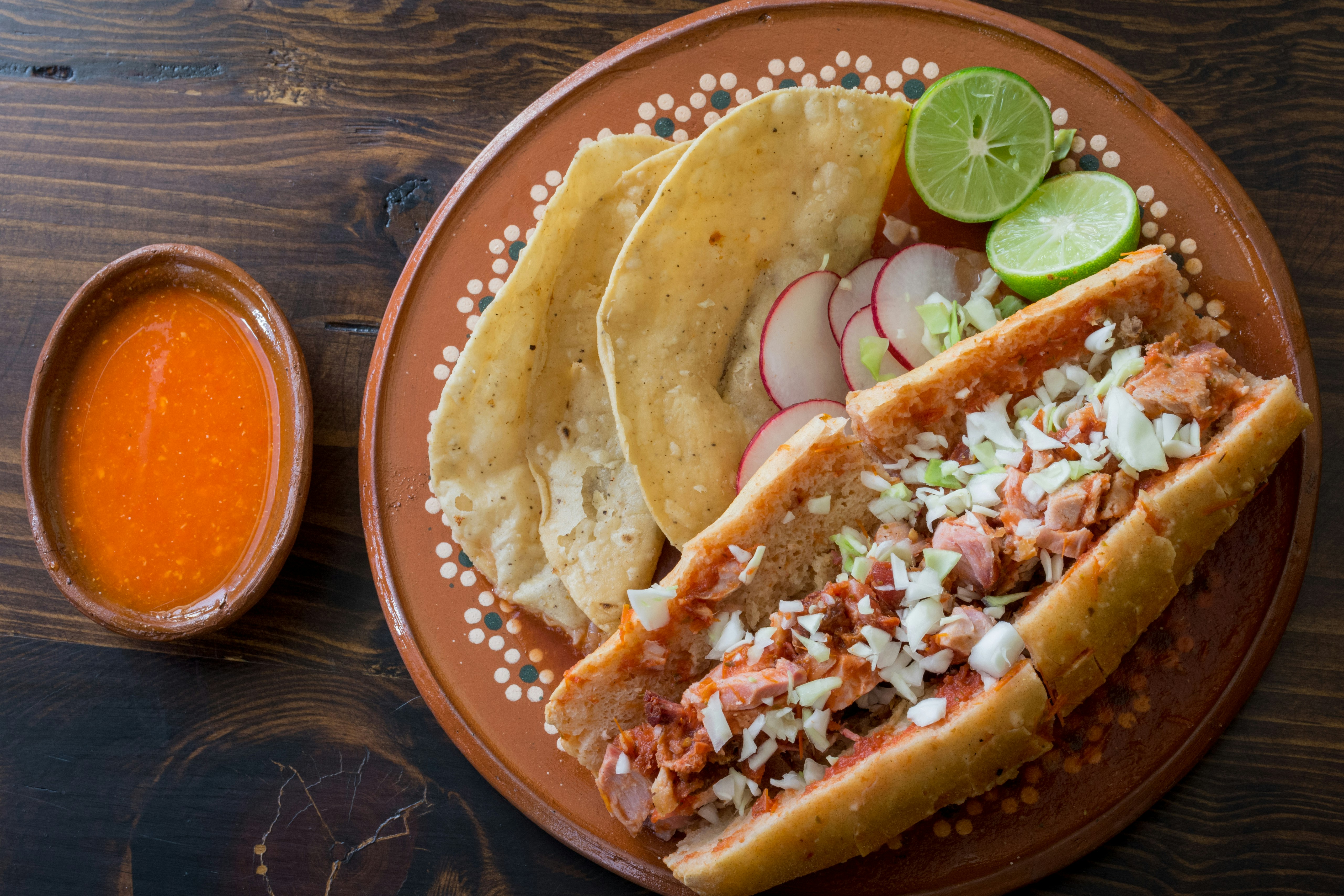 A tortas ahogadas sandwich, sitting in tomato-based sauce on a terracotta plate with tacos, and sliced limes.