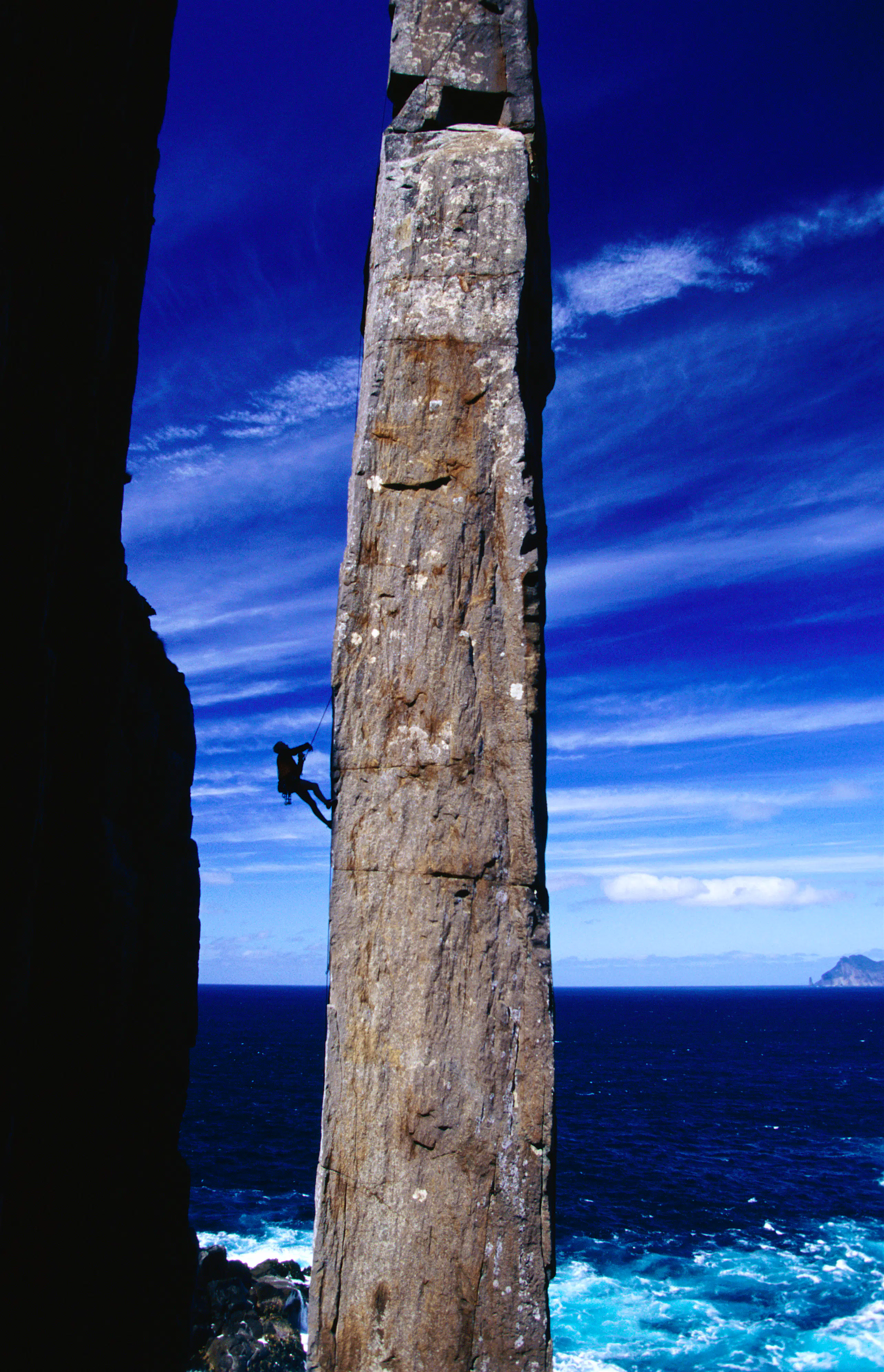 totem-pole-climb-tasmania-australia.jpg A narrow, rectangular spire of granite - known as the 'Totem Pole' rises up from the ocean into a blue sky streaked with wispy clouds; hanging from its side, and dwarfed by its size is a rock climber.