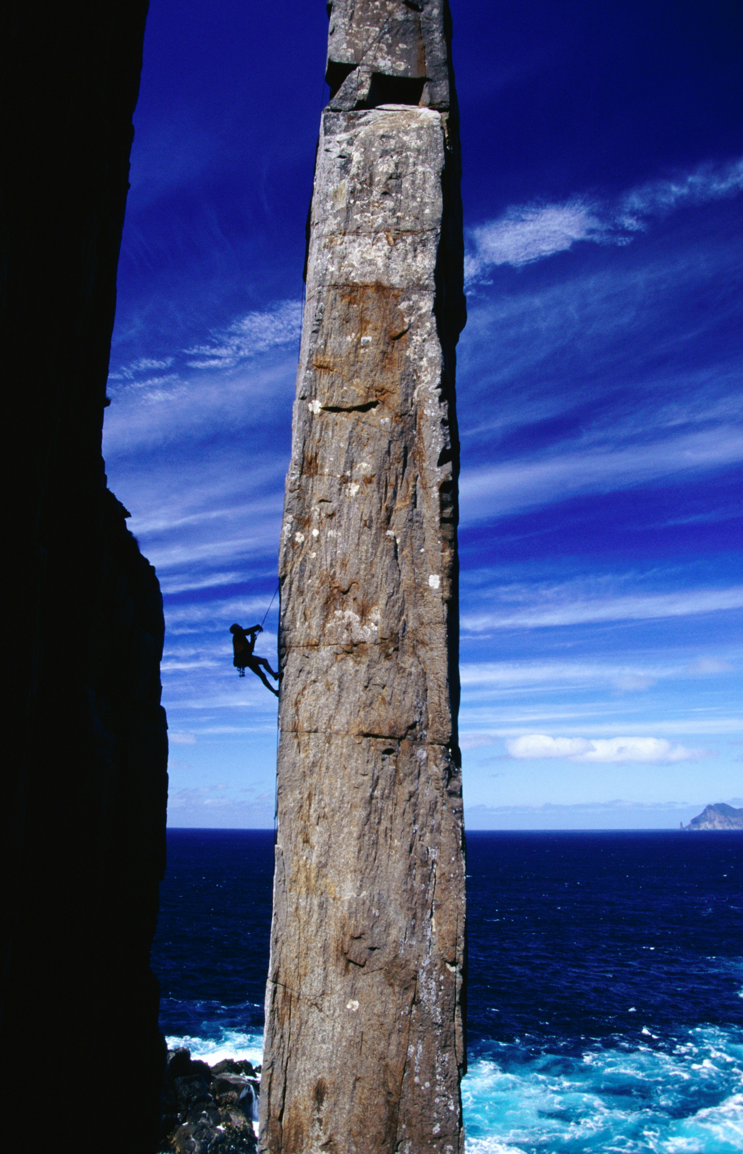 A narrow, rectangular spire of granite - known as the 'Totem Pole' rises up from the ocean into a blue sky streaked with wispy clouds; hanging from its side, and dwarfed by its size is a rock climber.