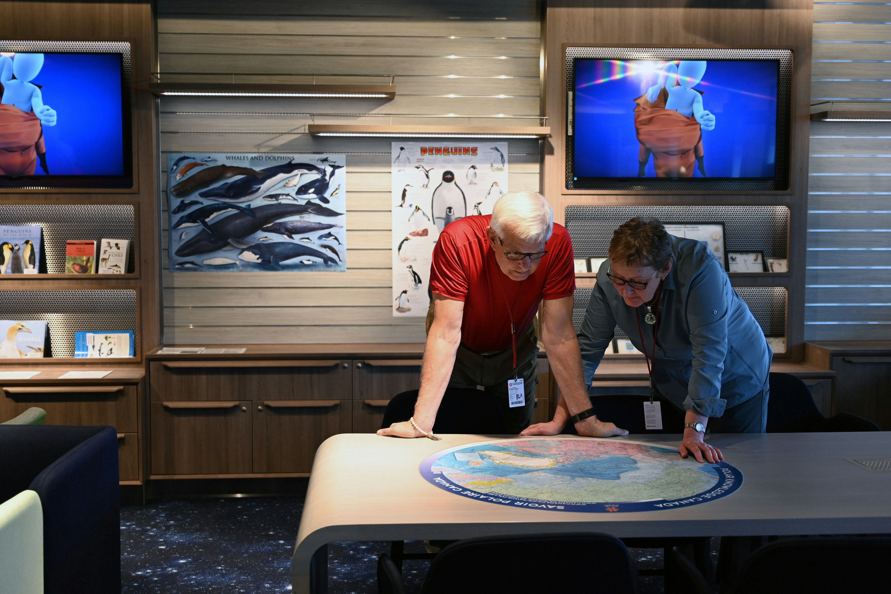A senior couple lean over a table in a library area aboard the MS Roald Amundsen; they're looking at a map of Antarctica.