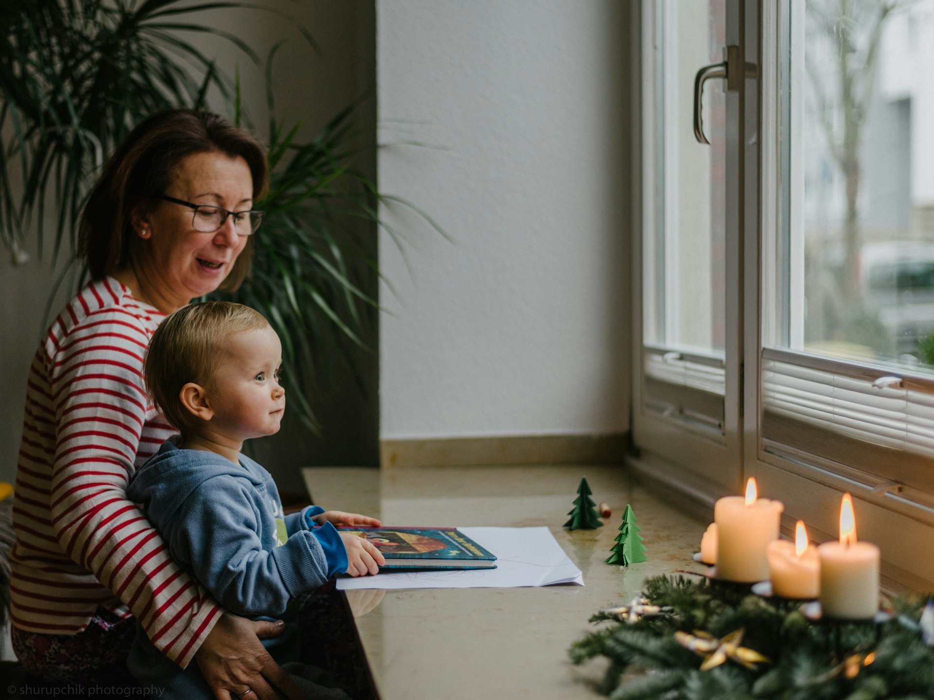 An older woman holds a toddler who looks out a window while holding a book. In the foreground is an advent wreath and paper Christmas trees