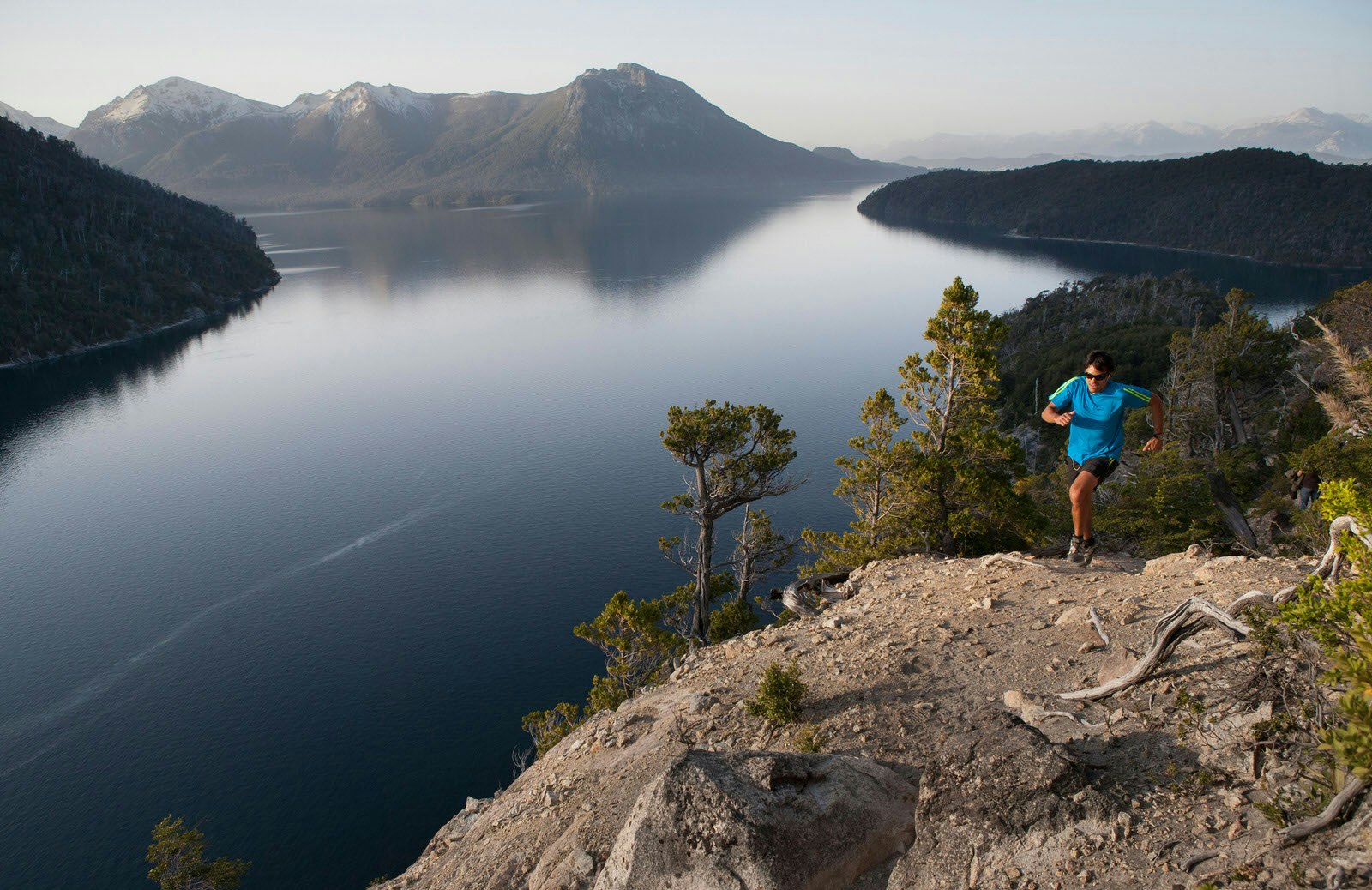 A runner ascends a rocky knoll, which slopes sharply down to a stunning lake; its shores are flanked by silhouetted, tree-covered sections of land, with mountains in the distance.