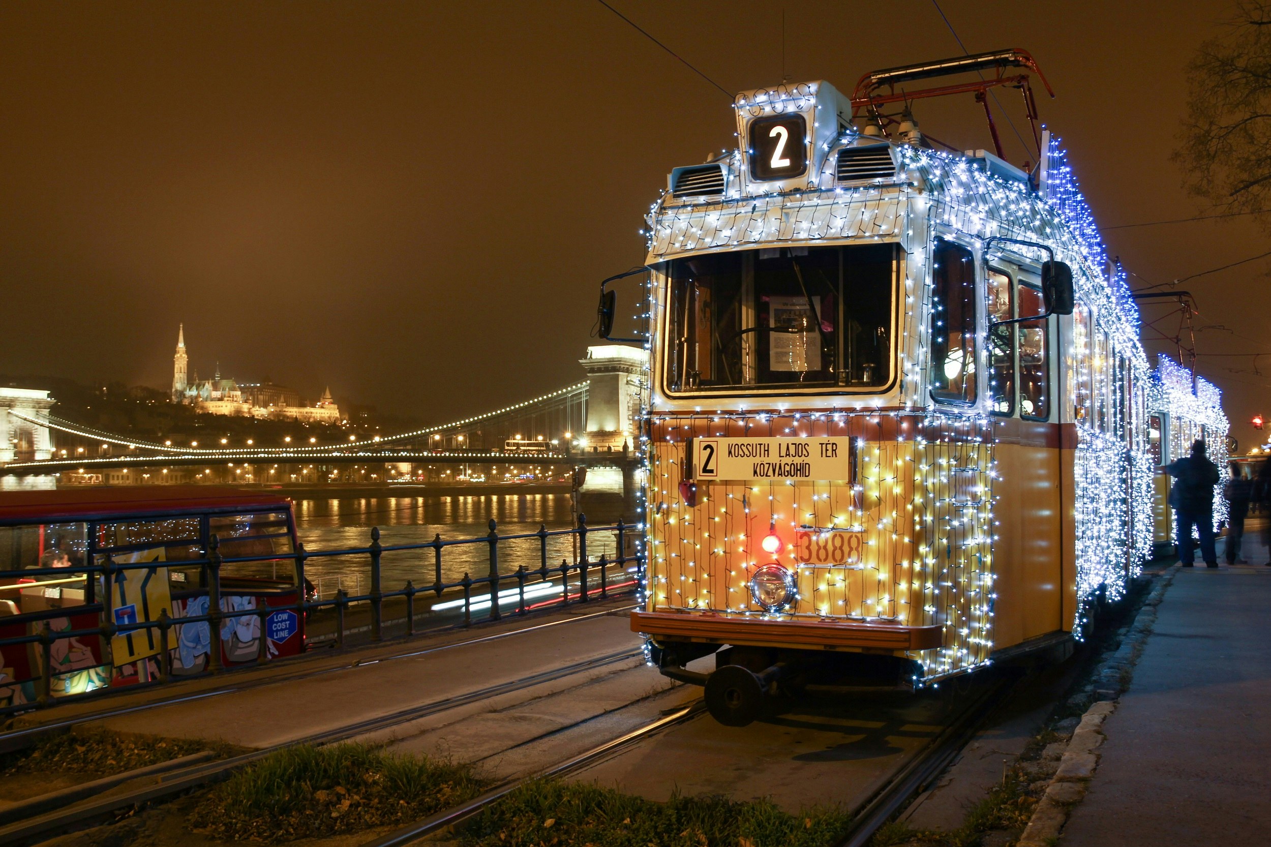 A shot from the front of a tram covered in small white and blue lights, stopped near the river