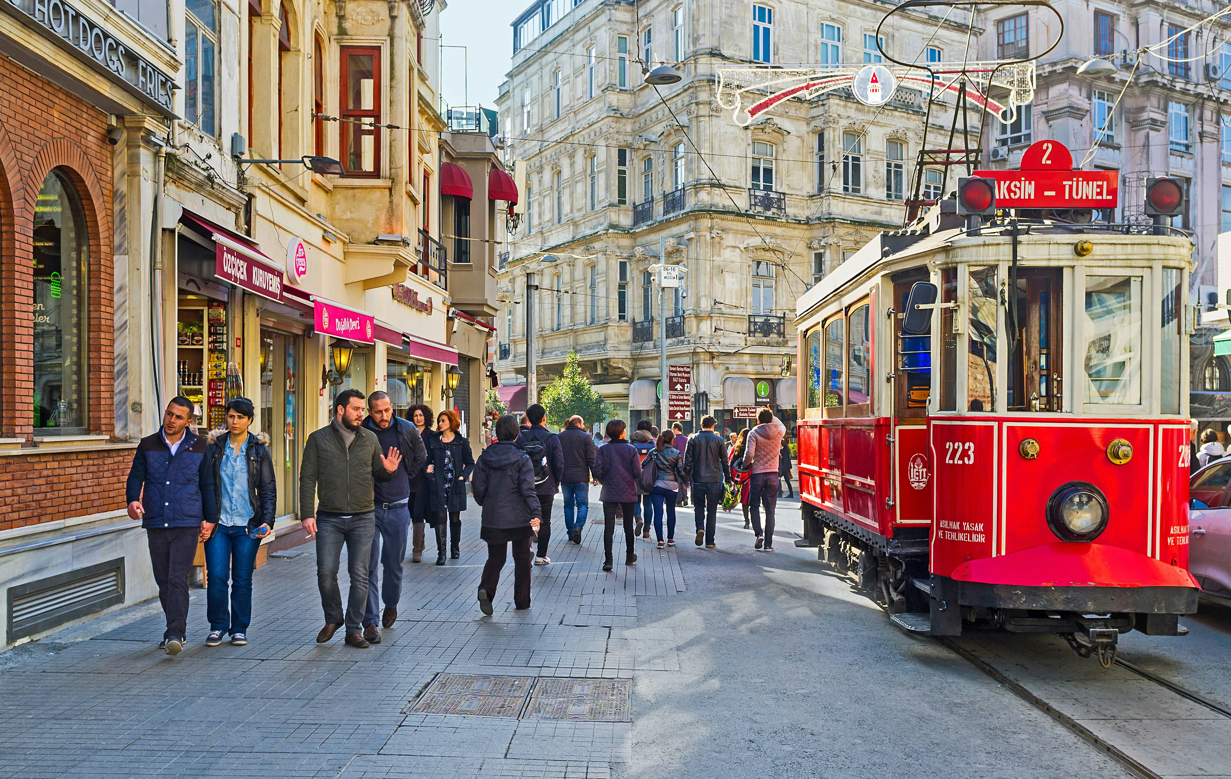 A bright red vintage tram is travelling down a pedestrian street; the pavement of the shopping street is busy with people.