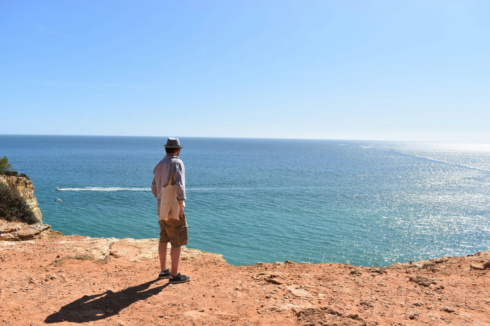 The author – wearing a shirt, shorts and hat and carrying a tote bag – looks out to a shimmering sea from a cliff the colour of burnt orange.