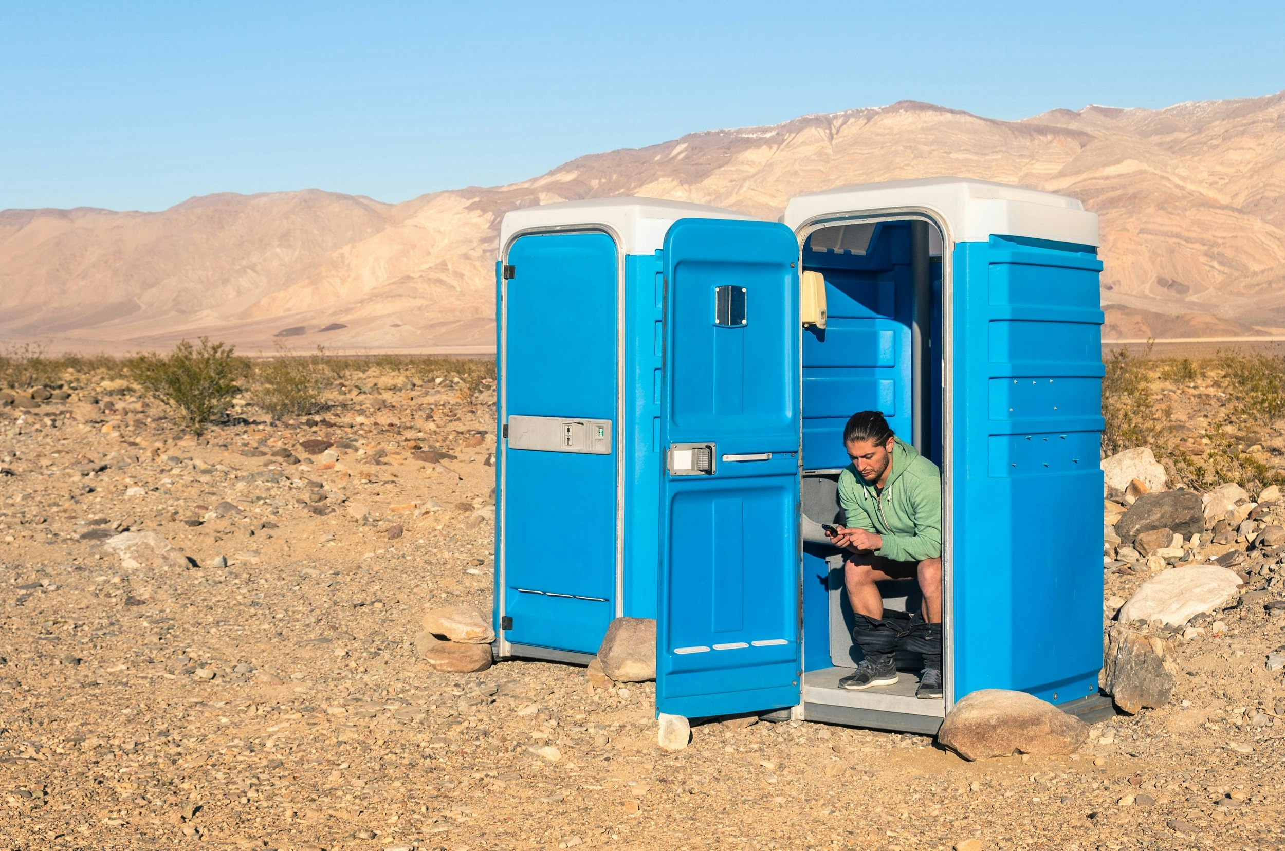 A man sits in a blue portable toilet with the door open; there is another loo next to his with the door closed. It's in Death Valley so that background is very barren.