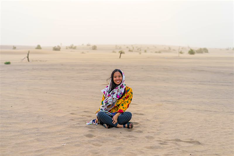 The author sits cross-legged in the desert, smiling, wearing a scarf over her hair 