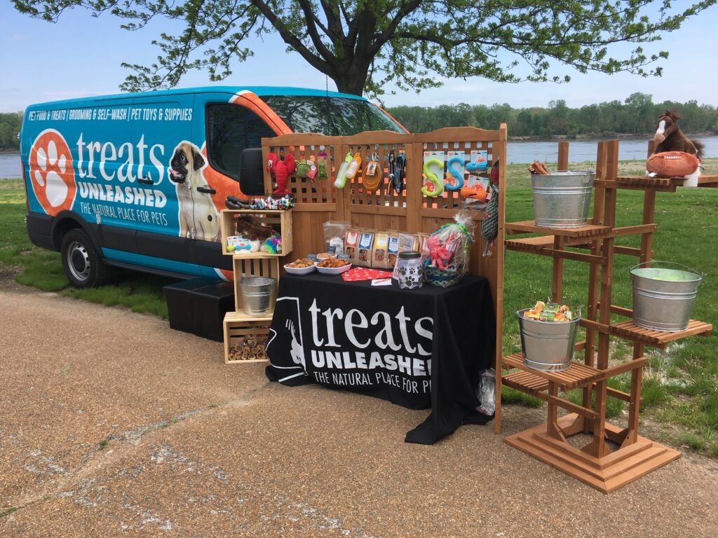 Wooden stands have been laid out with various dog treats beside a van covered with the Treats Unleashed company branding.